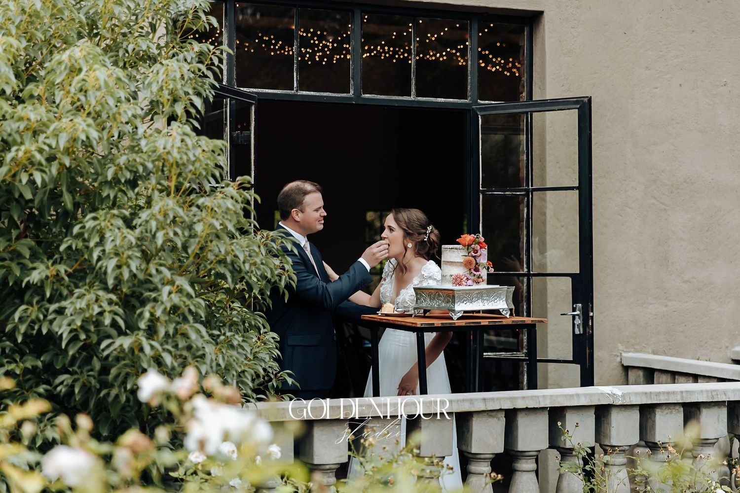 Bride and groom sharing an intimate moment at outdoor table with cake on a stone balcony.