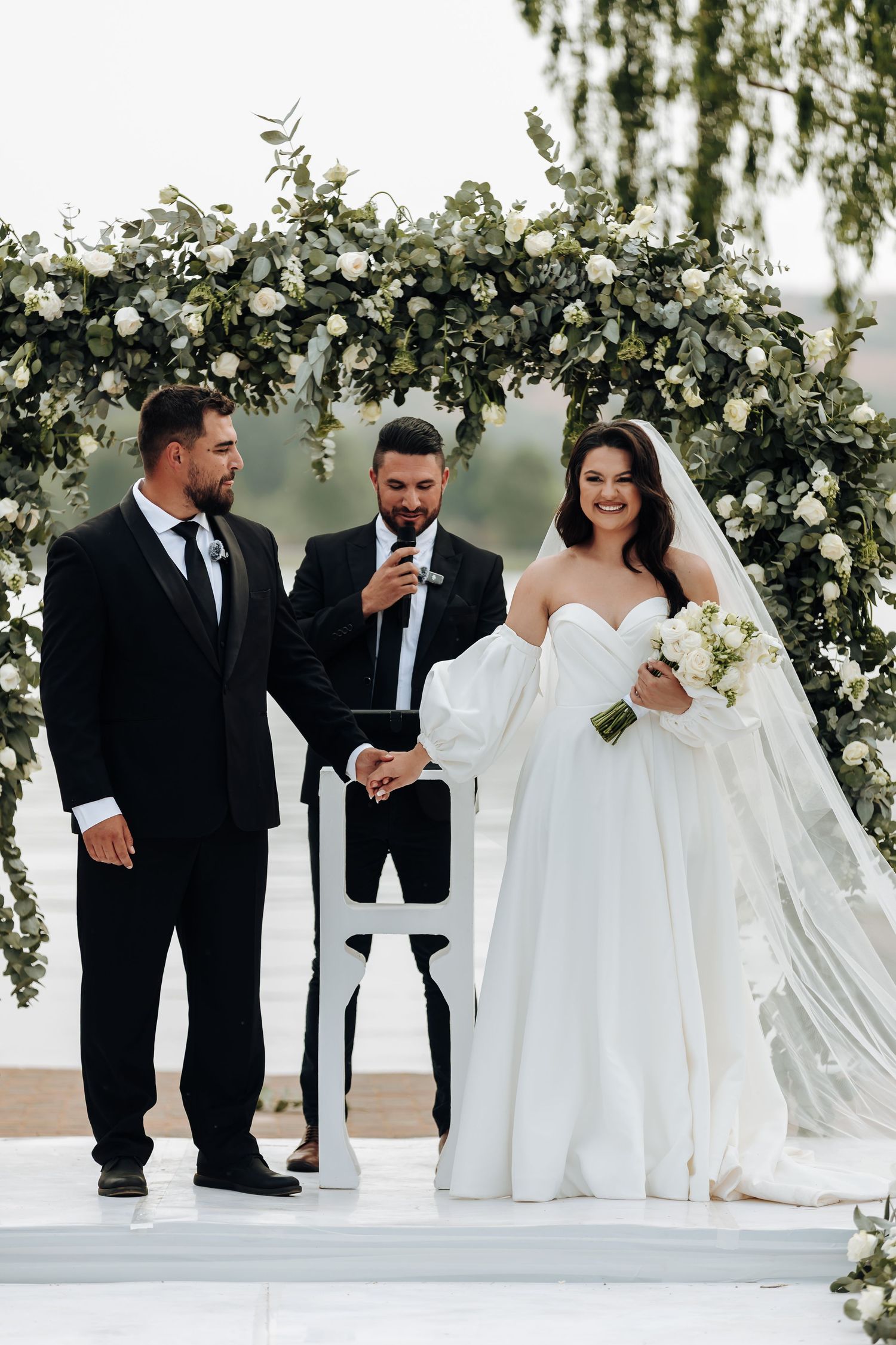 A wedding party in black and white attire poses under an elegant floral arch during an outdoor ceremony.