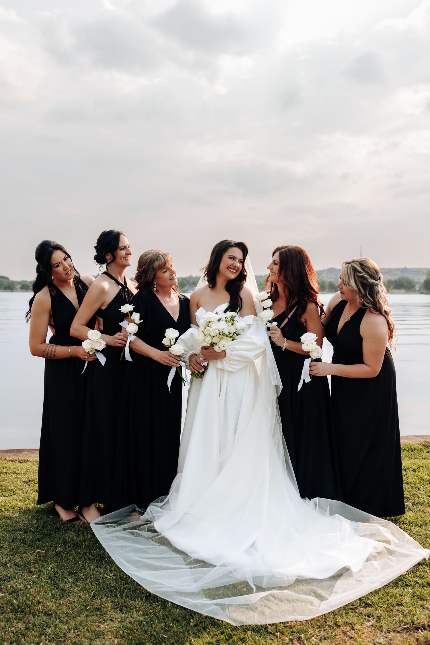 Wedding party in black dresses and white gown pose together laughing joyfully on grassy lawn by waterfront.