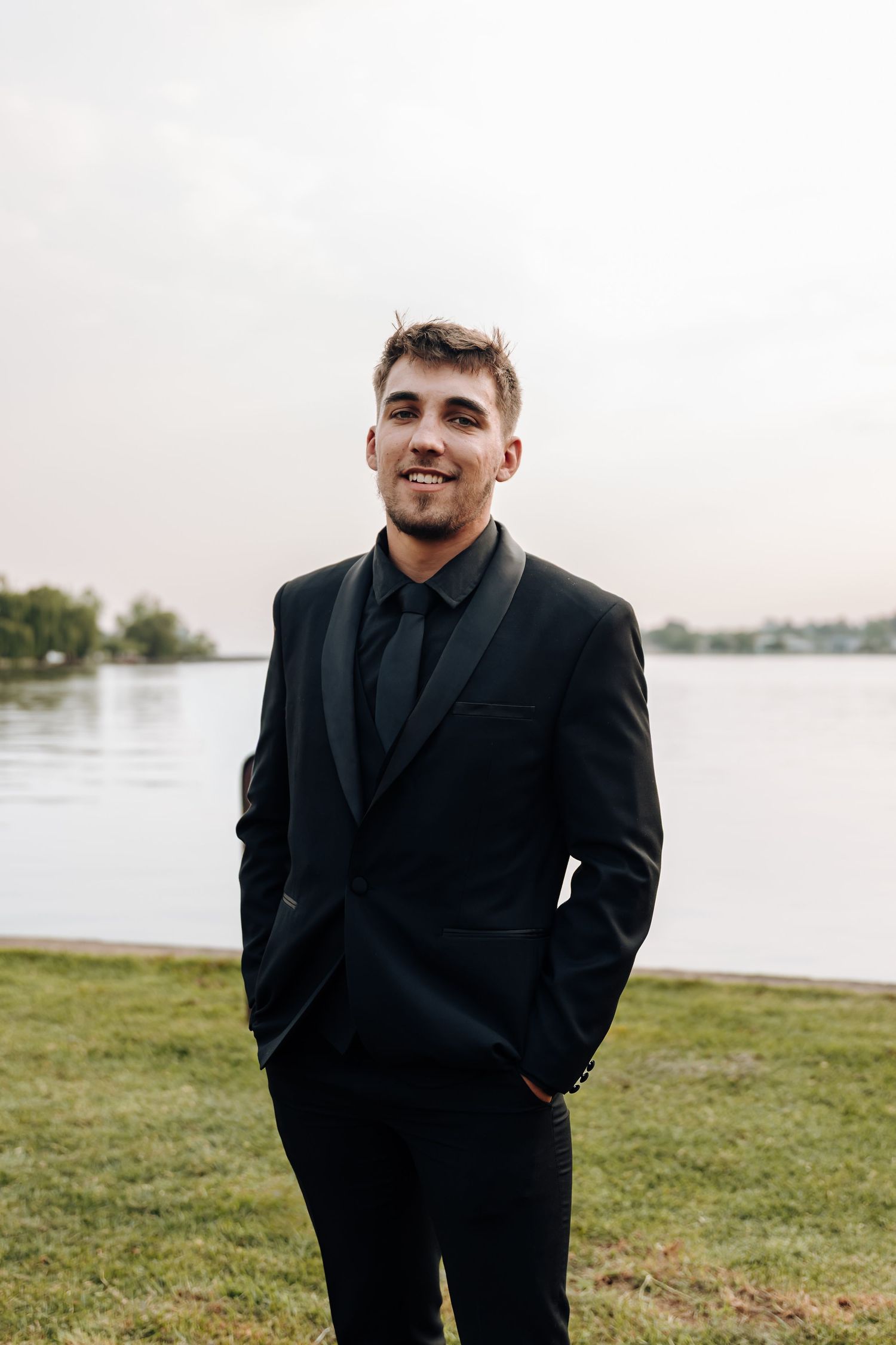 A person in a black suit stands outdoors near a waterfront on a grassy area in overcast weather.