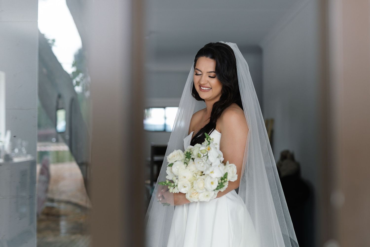 A bride in a white wedding gown holds a bouquet of white flowers while posing in an elegant indoor setting.