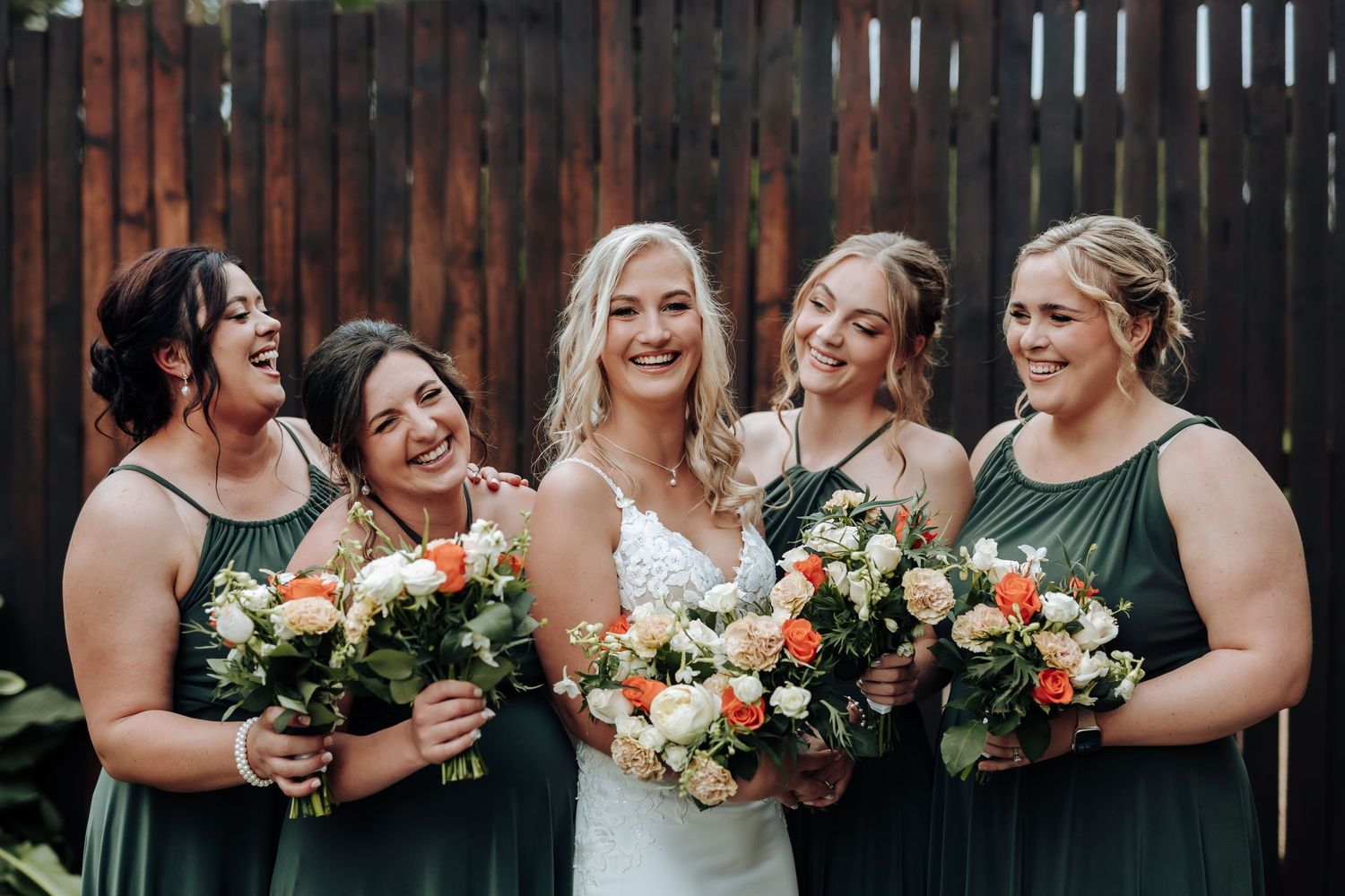 A bridal party in sage green dresses holds peach and cream floral bouquets while laughing together against a wooden fence.
