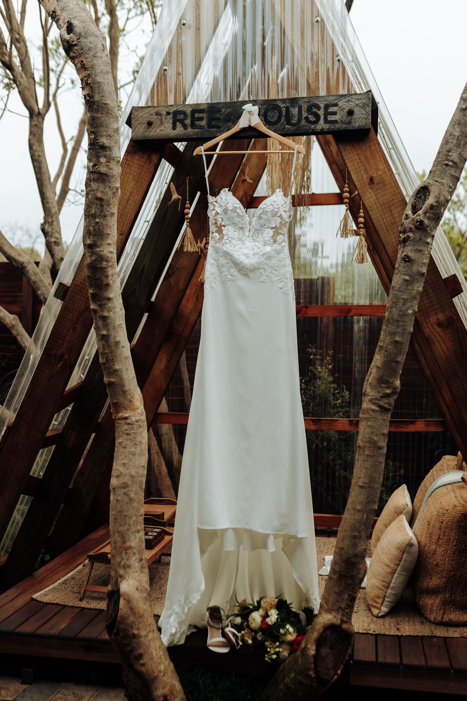 A white wedding dress hangs elegantly from a birch tree branch outside a rustic treehouse venue.
