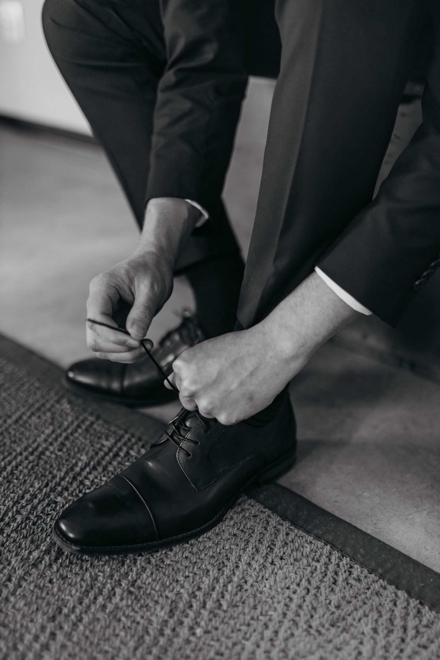 Close-up view of hands tying black leather dress shoes on carpeted floor in black and white photography.
