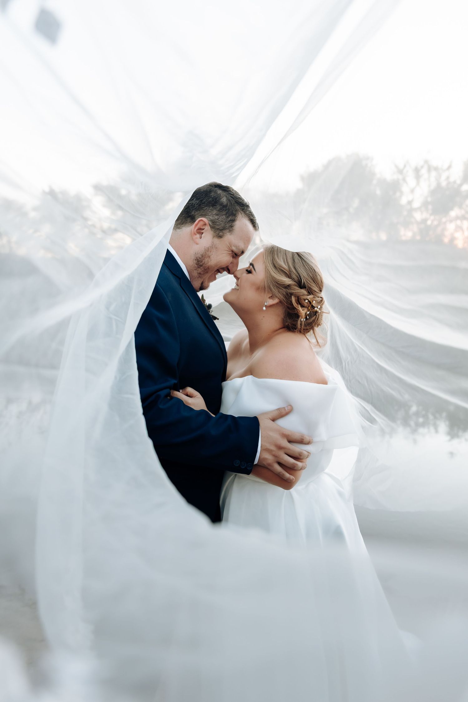 Bride and groom share a romantic forehead touch wrapped in a flowing wedding veil outdoors.