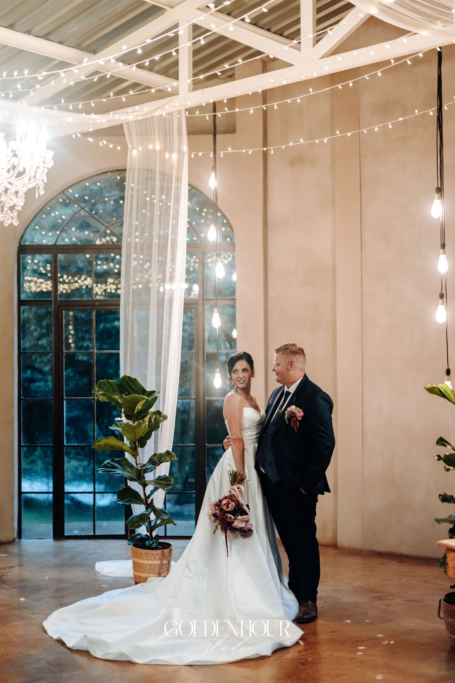 Bride and groom standing together in an elegant venue with fairy lights, sheer drapes, and arched windows.