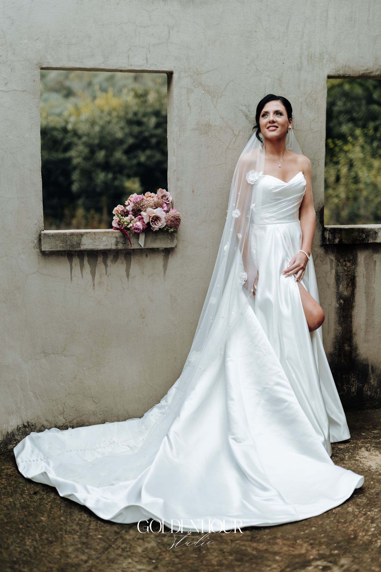 Bride in strapless satin ballgown with long train and veil holds pink bouquet outdoors at rustic stone venue.