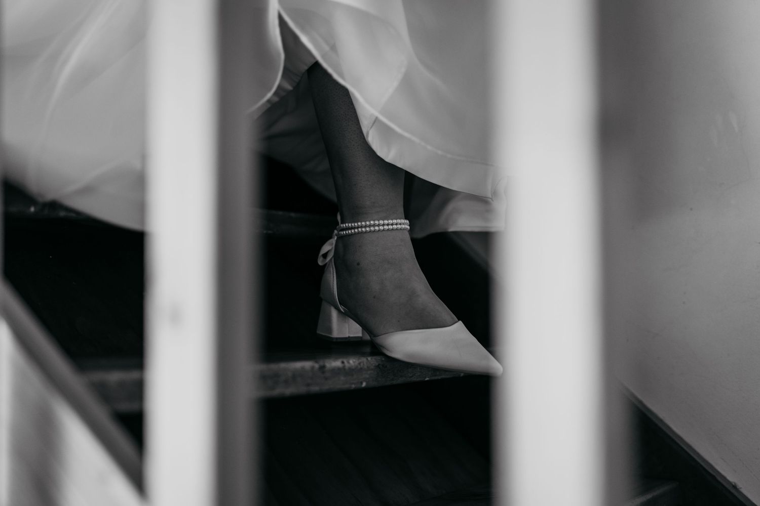 Black and white photo of a bride's heeled shoes with ankle strap on staircase, wedding dress visible above.