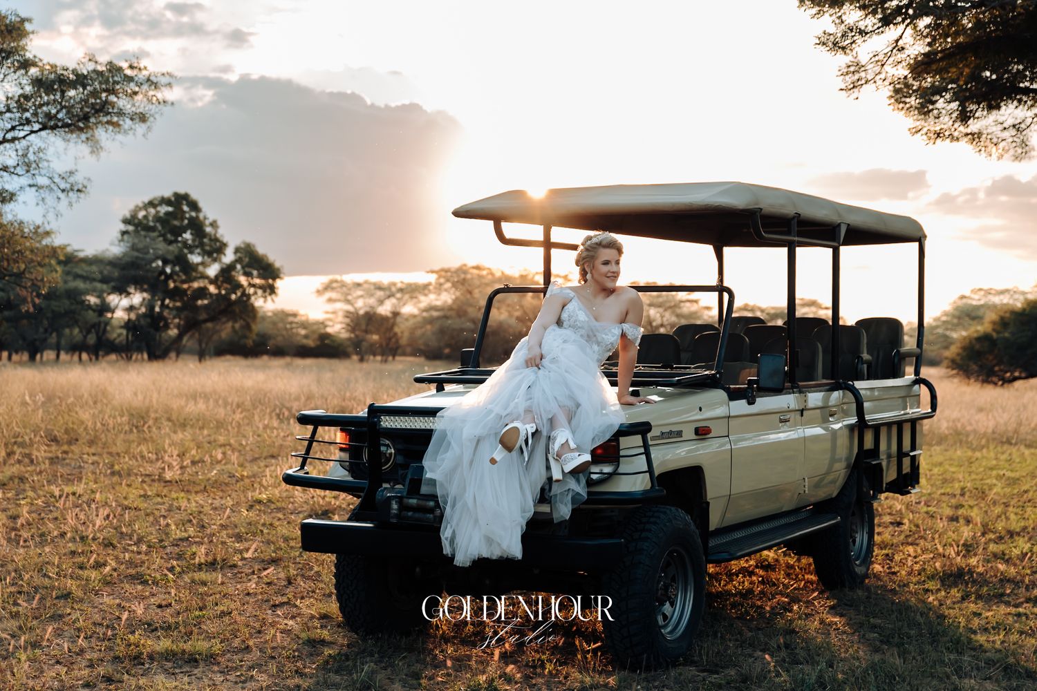 Bride in white gown poses elegantly on safari jeep at golden sunset in African savanna.