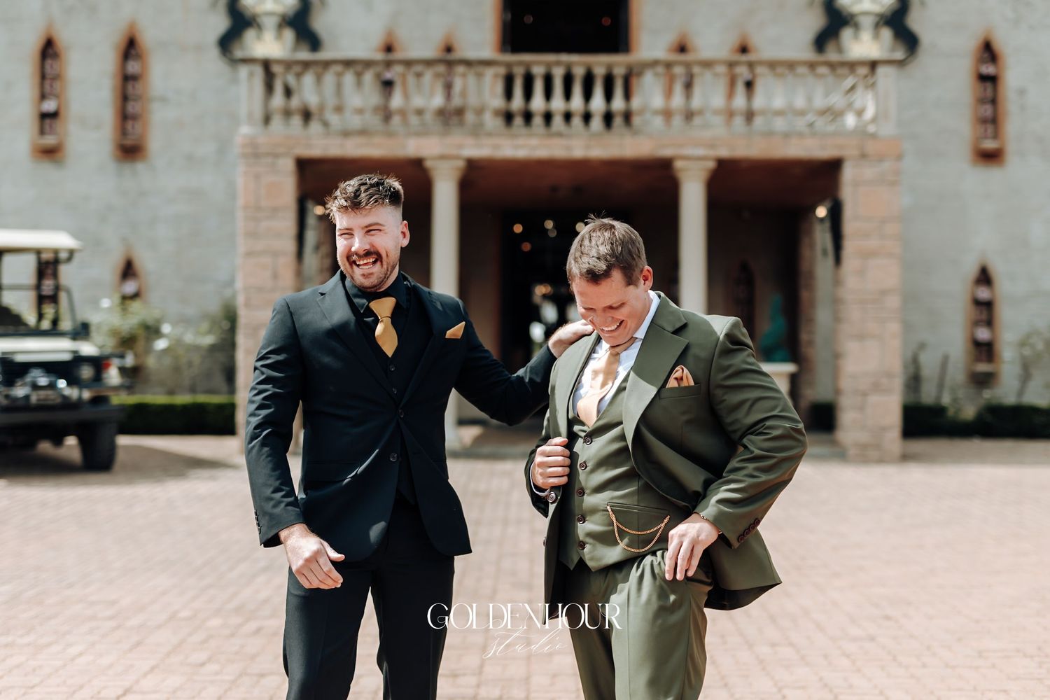 Two men in elegant wedding suits, one in black and one in olive green, laughing outside luxury venue.
