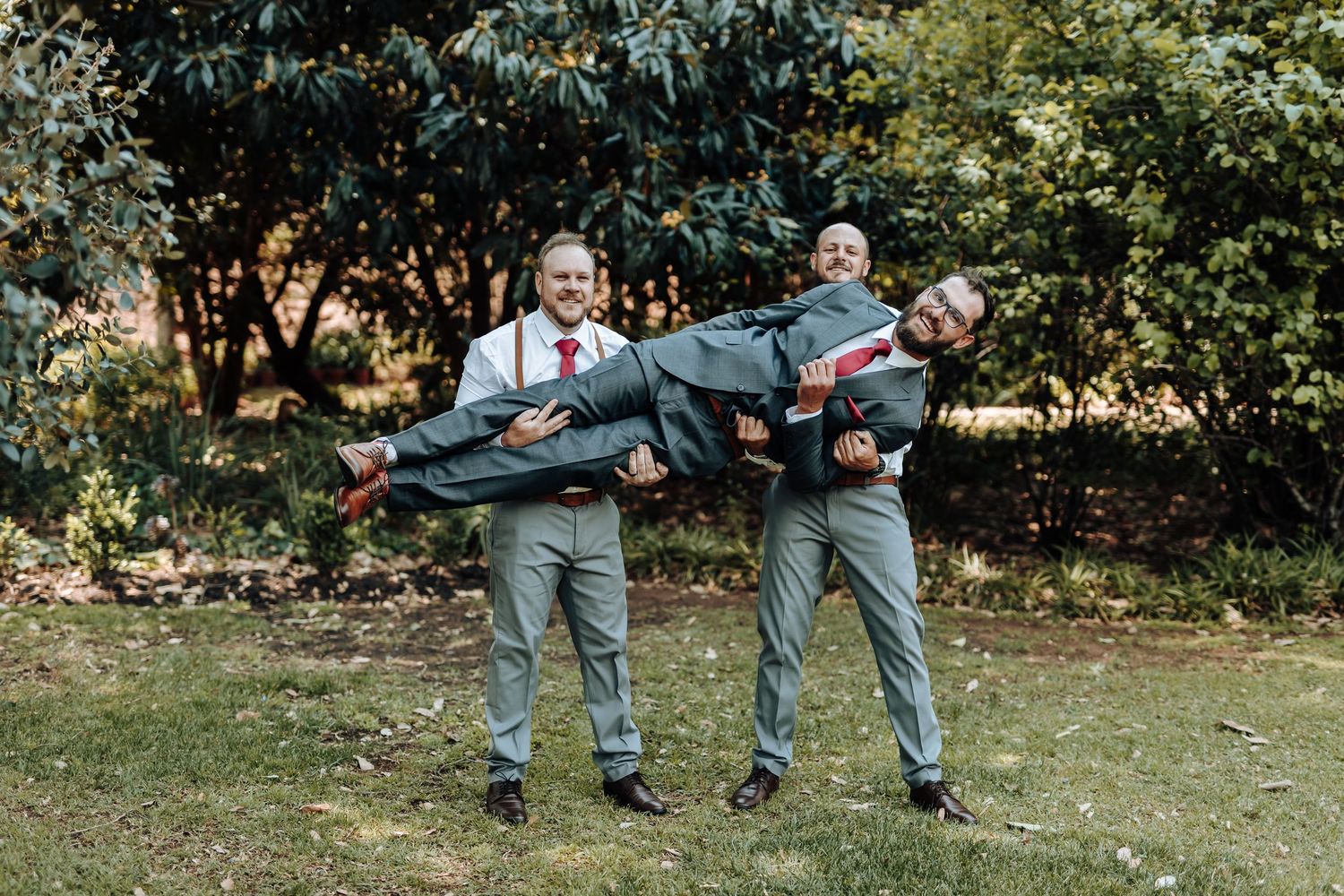 Two groomsmen in gray suits playfully carrying their friend horizontally for a fun photo.