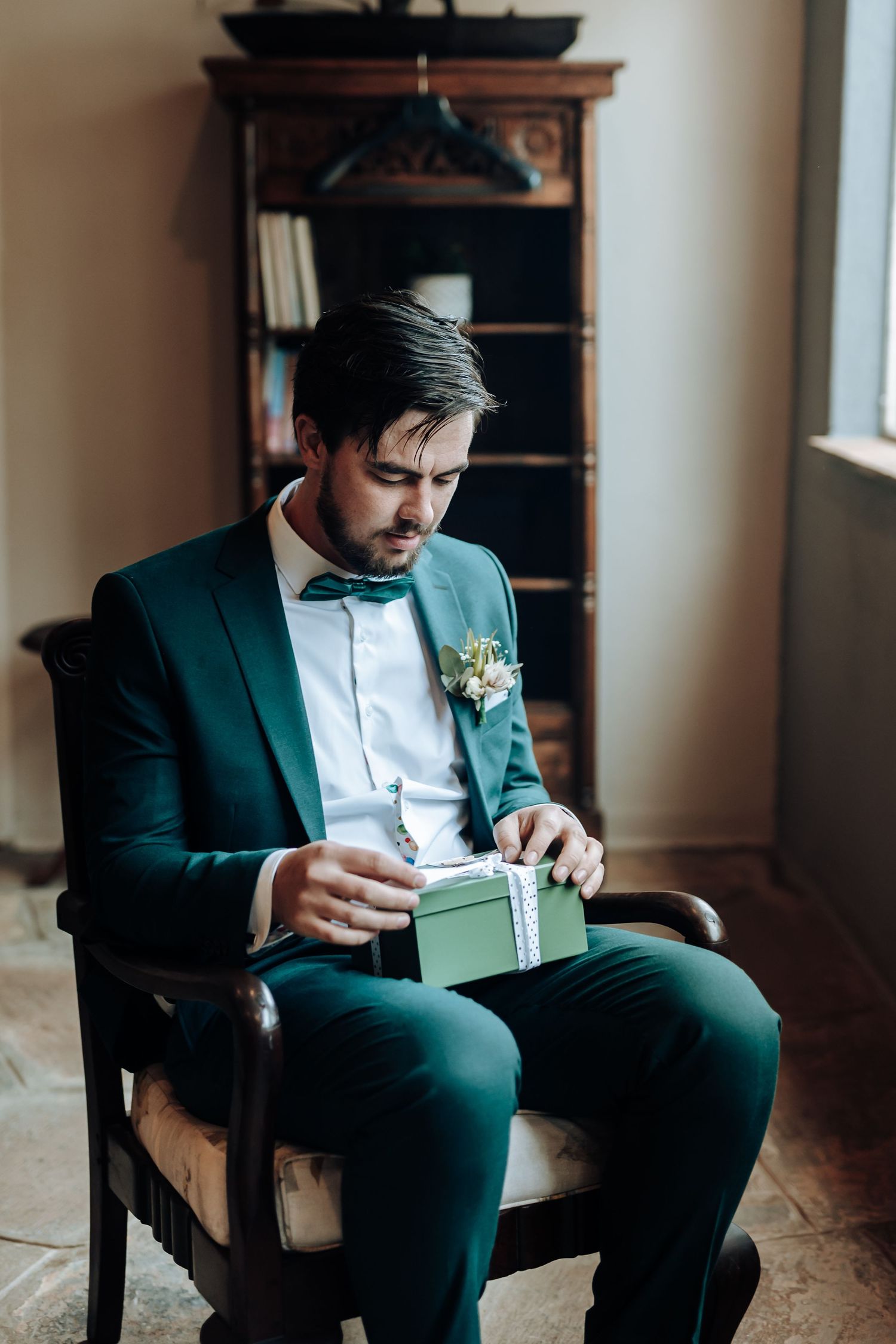 Groom in emerald suit opens gift while seated in vintage chair by window.