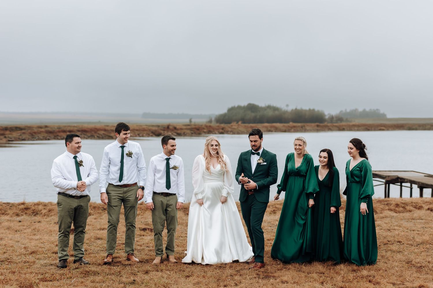 Wedding party in emerald dresses and white shirts poses by lakeside dock.