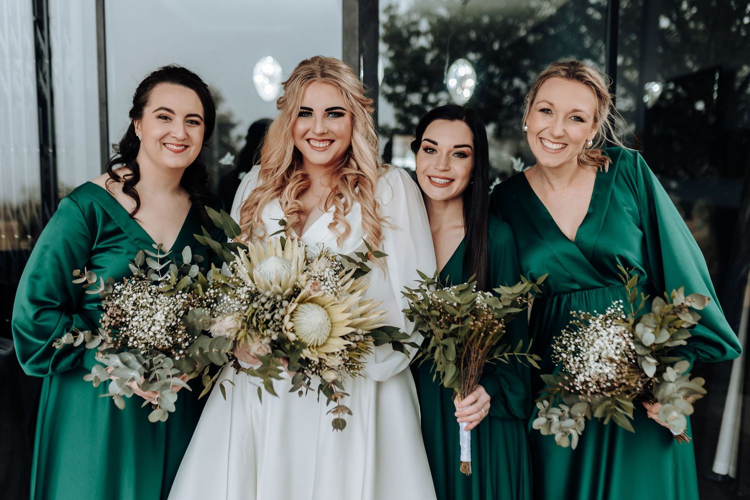 Bride in white gown poses with bridesmaids wearing emerald green dresses holding matching bouquets.