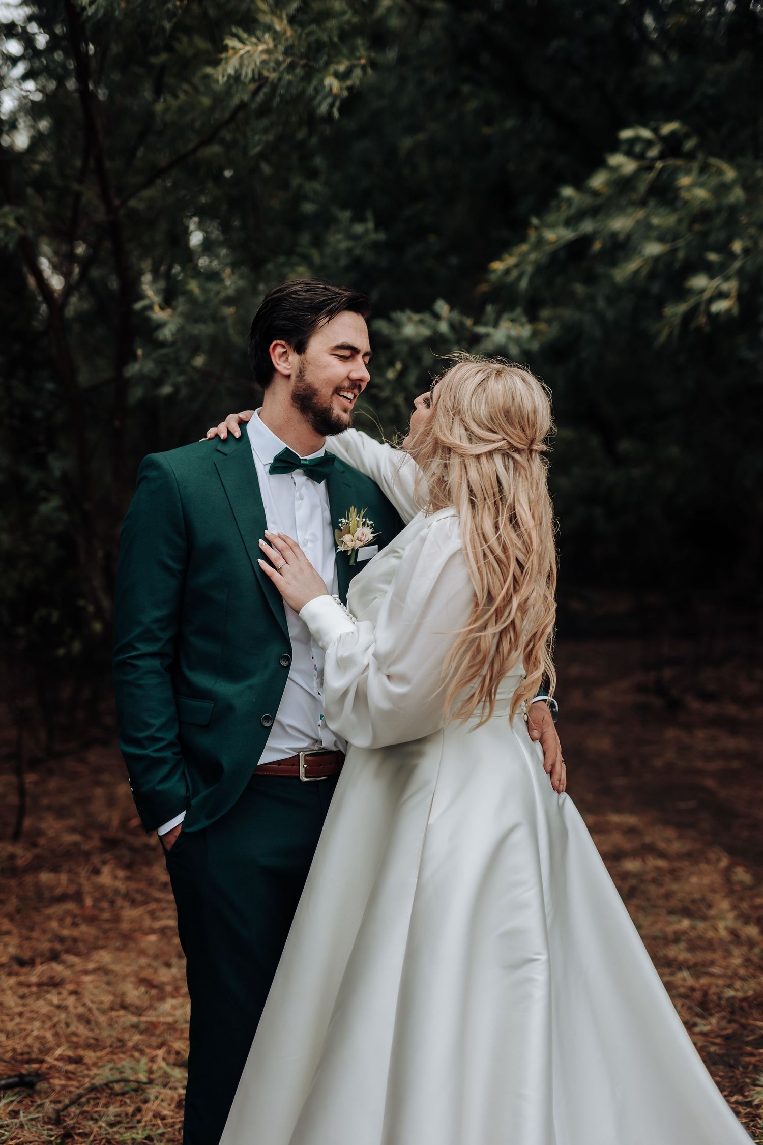 Newlyweds share tender moment in wooded setting, bride in elegant gown and groom in emerald suit.