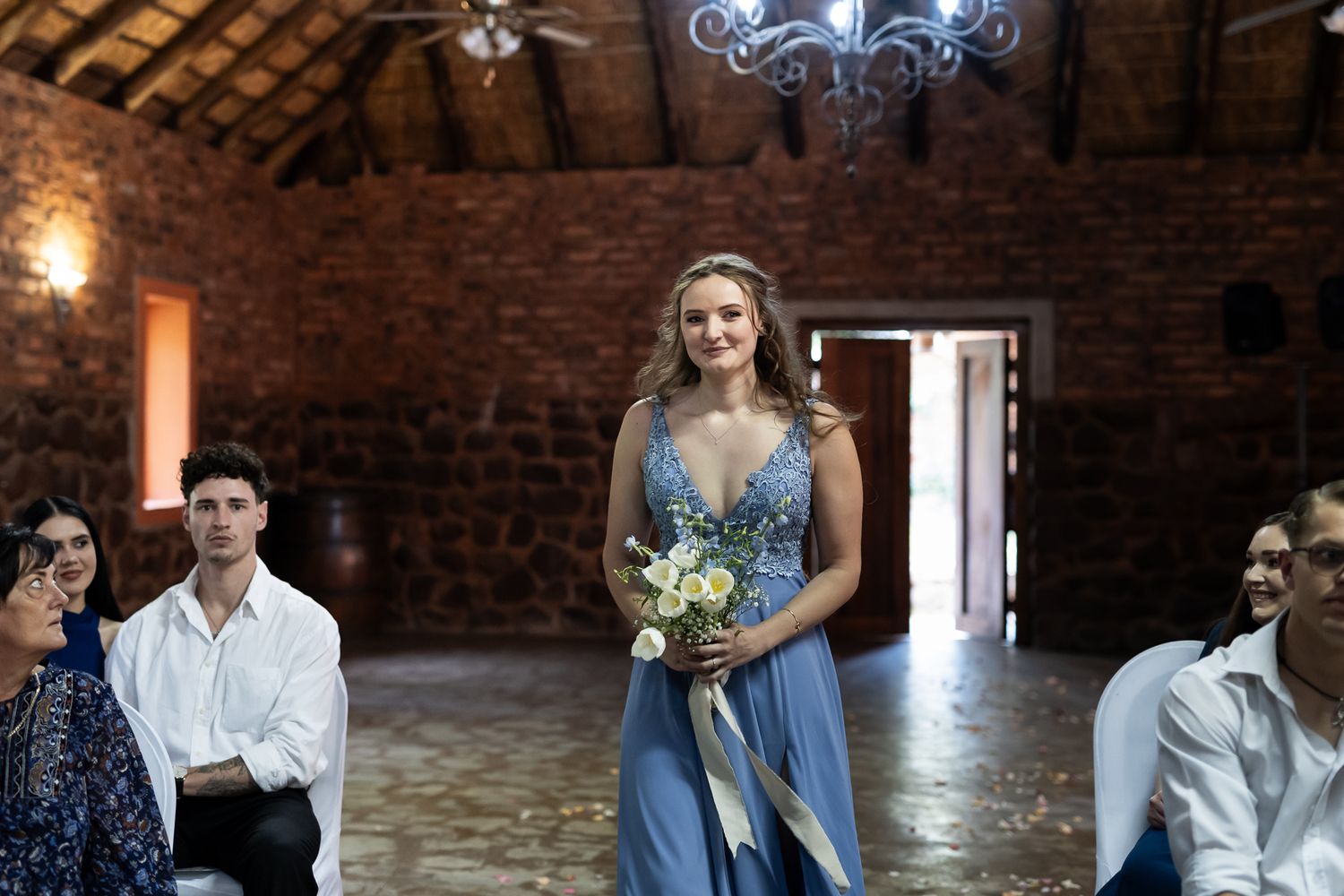 Wedding party member poses in blue dress holding bouquet inside rustic brick venue with chandelier.
