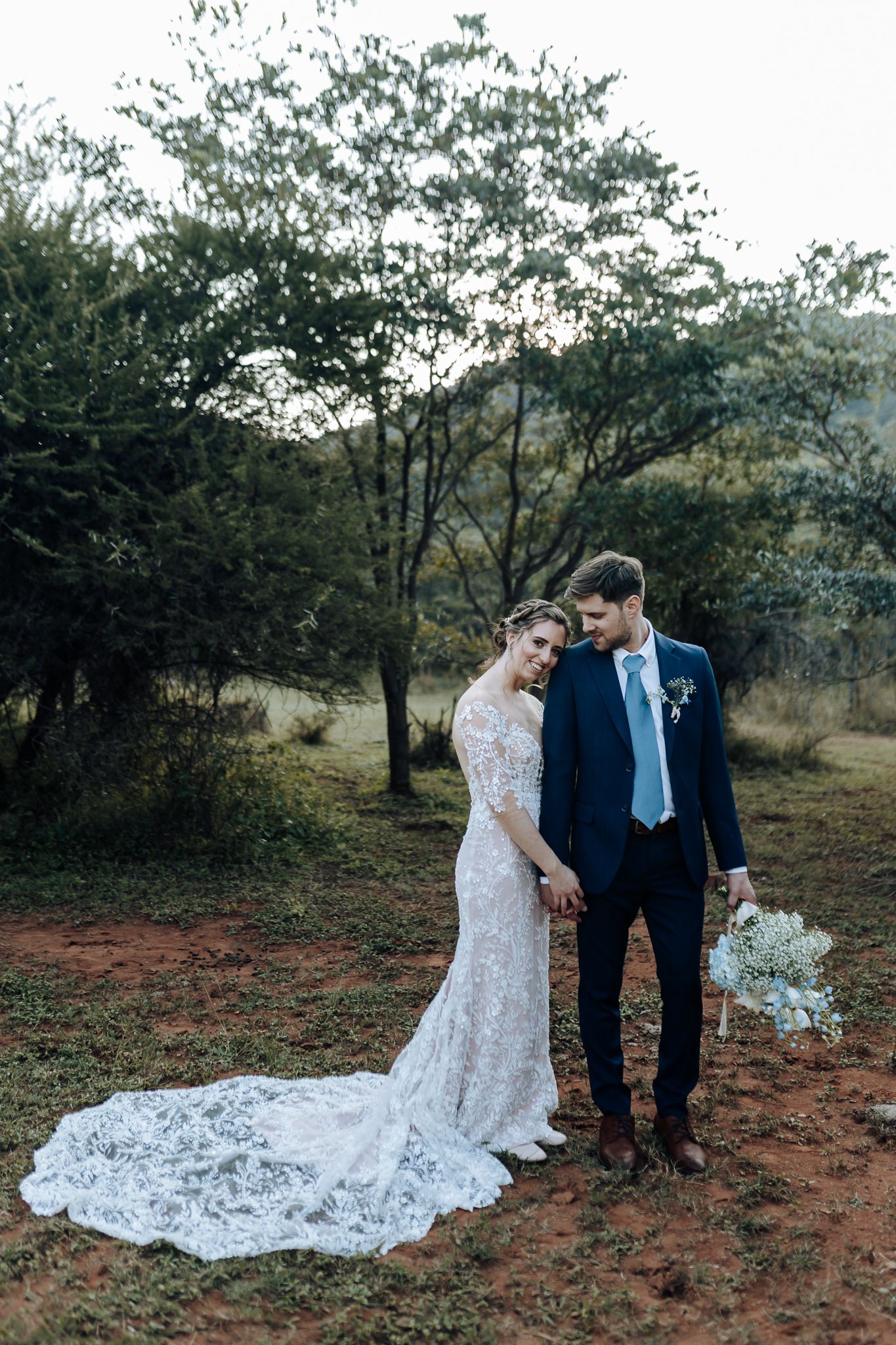 Full-length wedding portrait showcasing bride's dramatic lace train against natural outdoor setting.