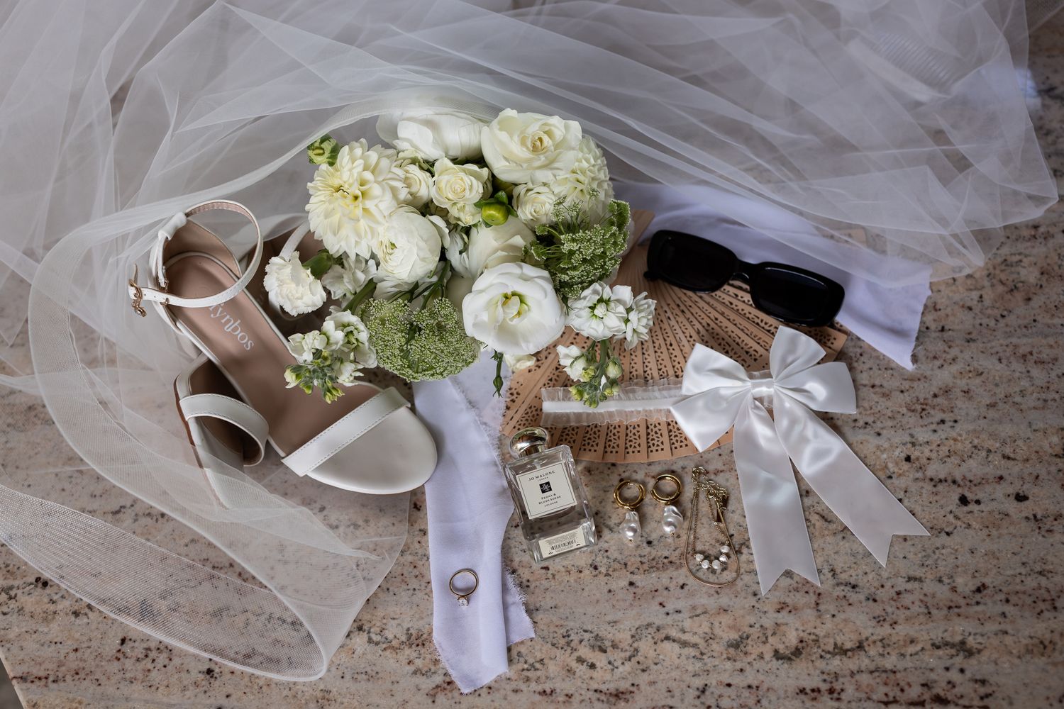Wedding details featuring white bouquet, shoes, and accessories arranged on textured surface.