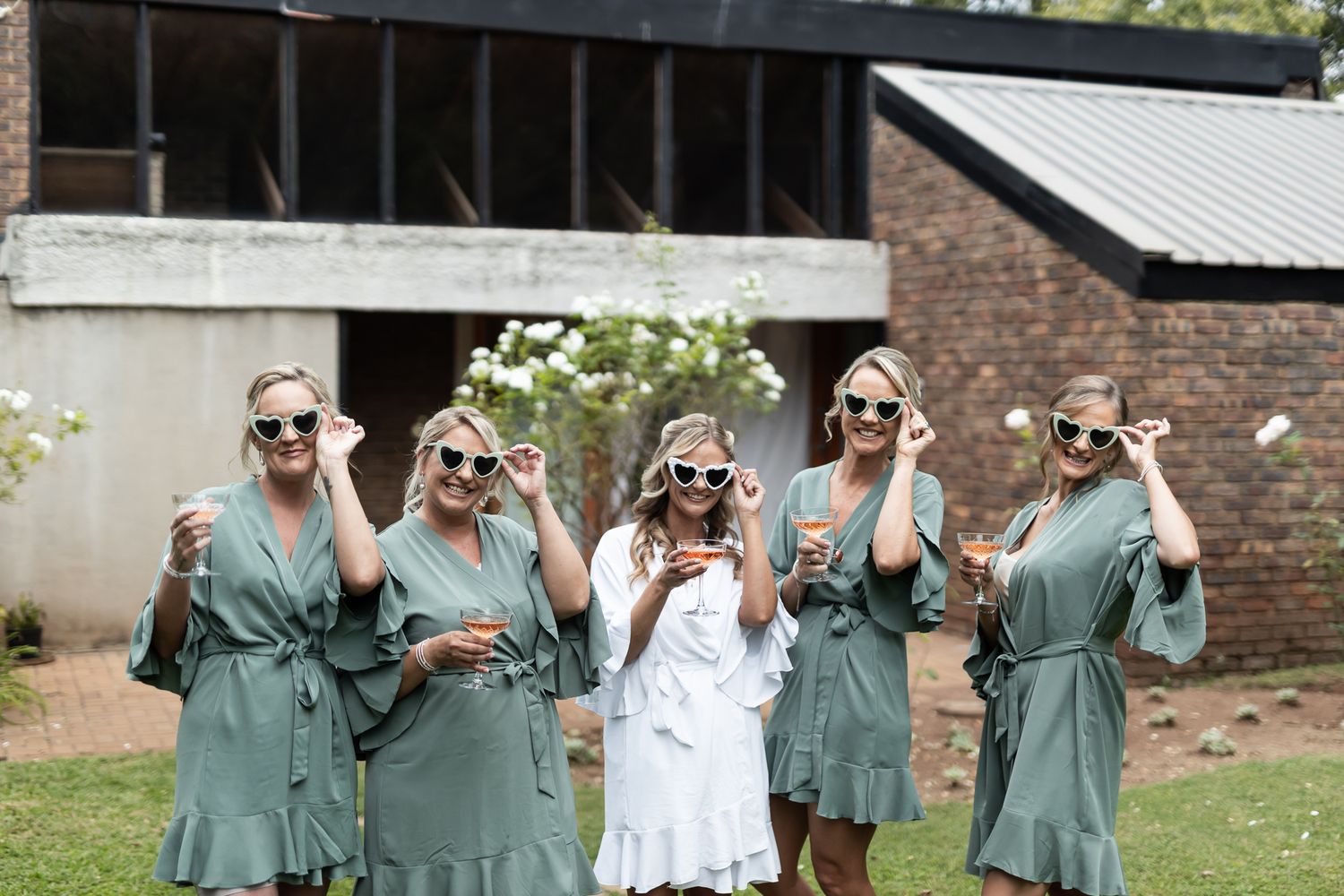 Bridesmaids in matching green robes and sunglasses pose with the bride on a sunny lawn.