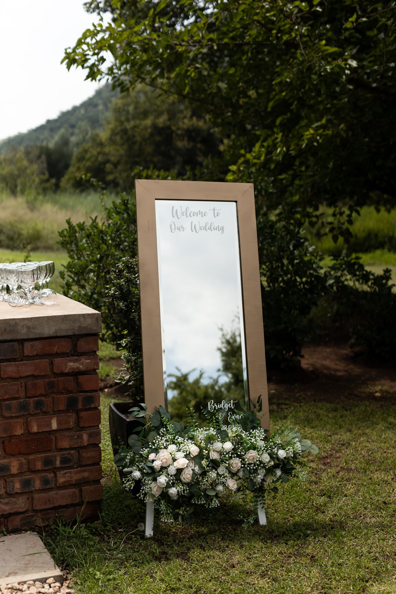 Elegant mirror welcome sign decorated with white floral arrangements in outdoor garden setting.