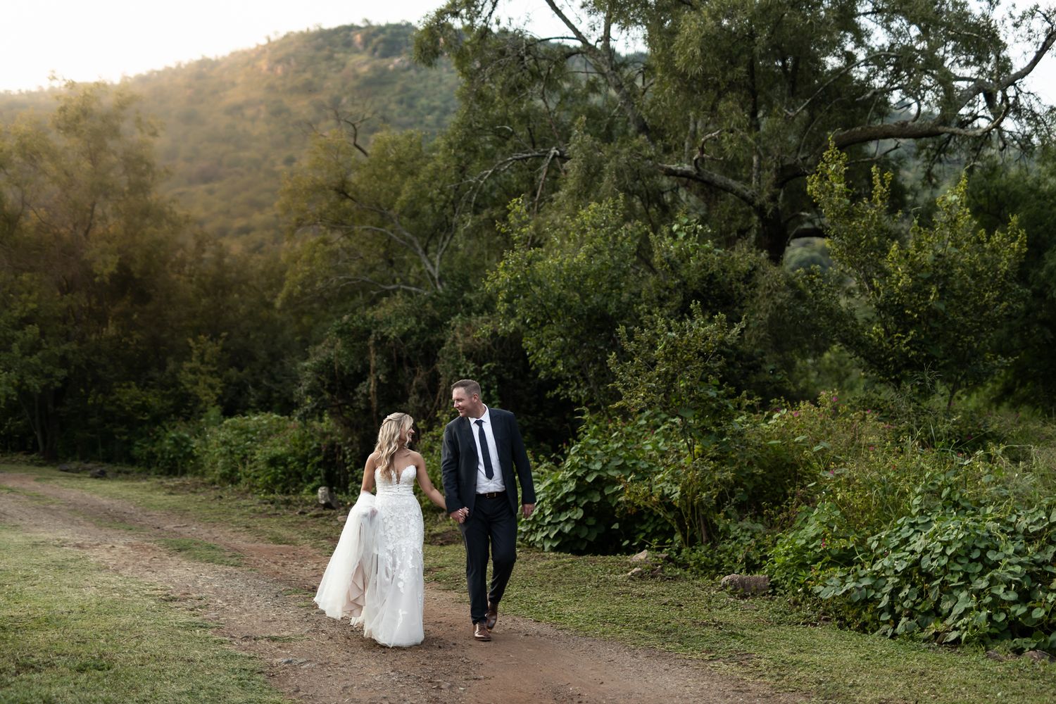 Wedding couple strolls together on a natural path with golden sunlight filtering through trees.