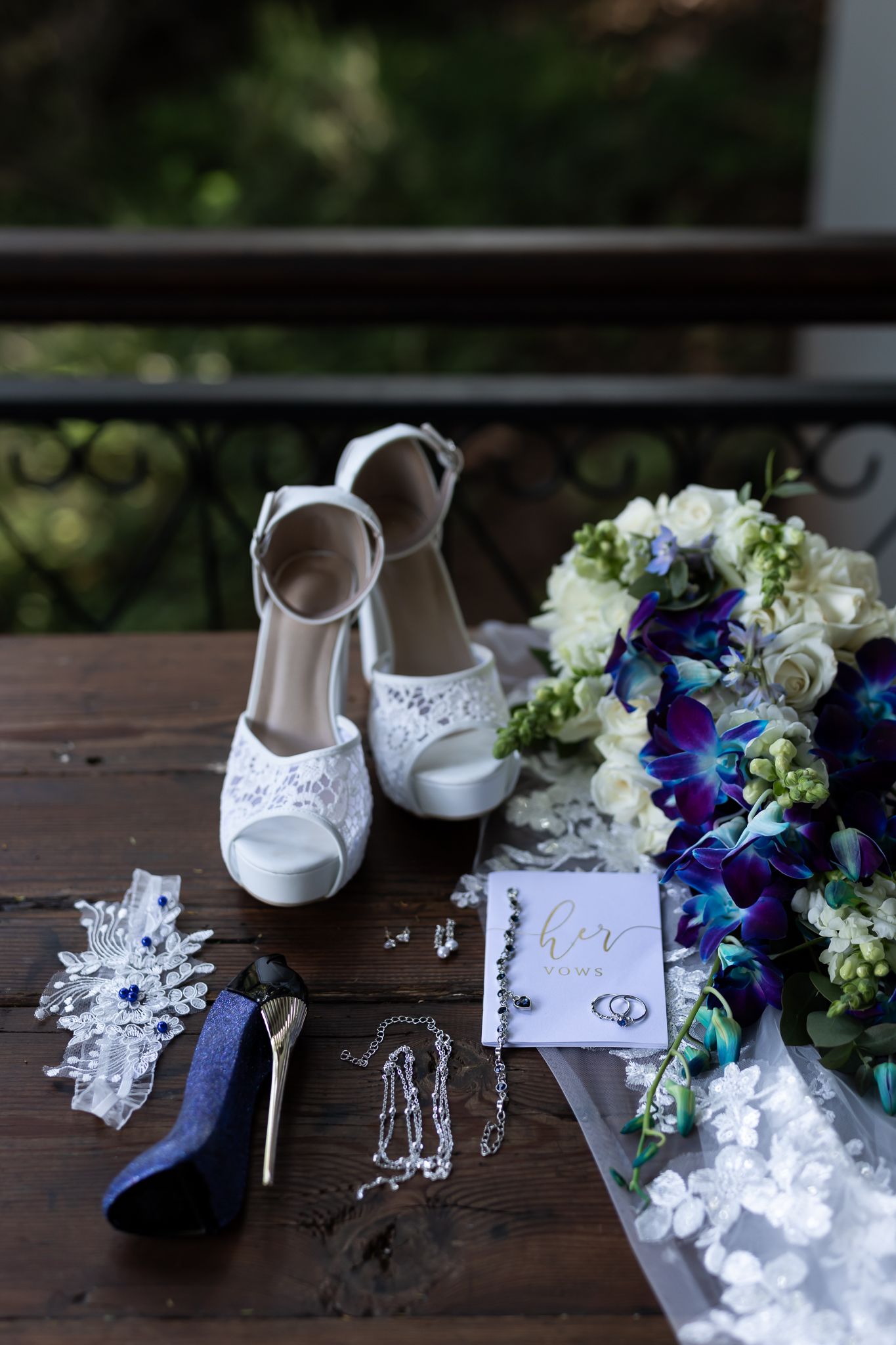 Wedding accessories including white lace shoes, blue and white floral bouquet, and jewelry arranged on wooden surface.
