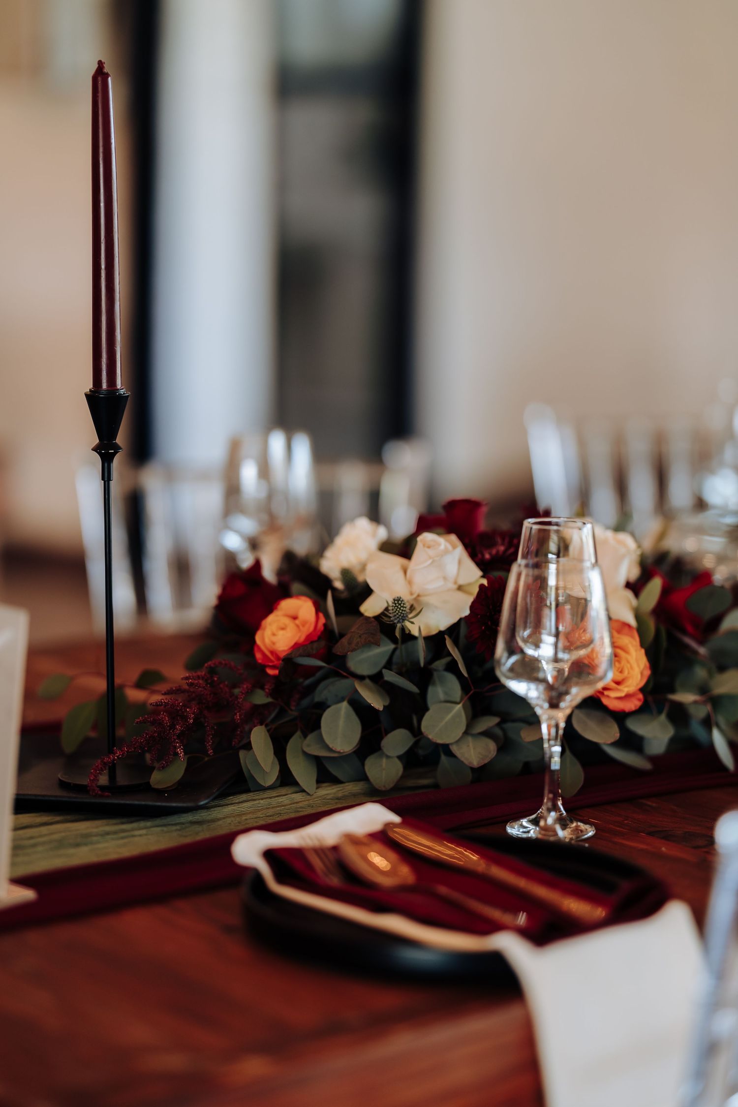 Elegant wedding table setting with red roses, eucalyptus, black candlesticks, wine glasses, and gold cutlery on a wooden table.