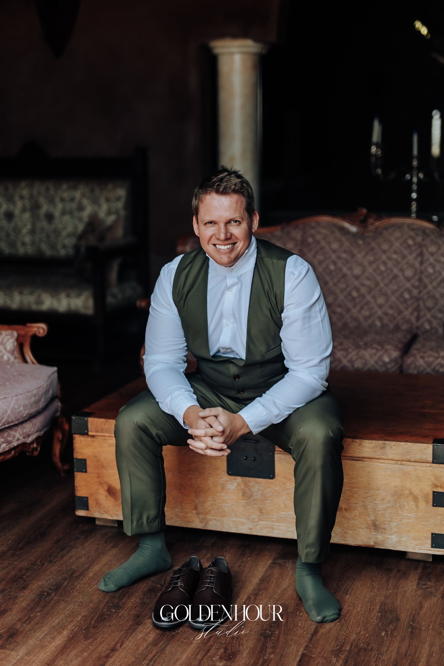 Smiling groom in olive green vest and trousers sits on wooden chest holding hands together at wedding venue.