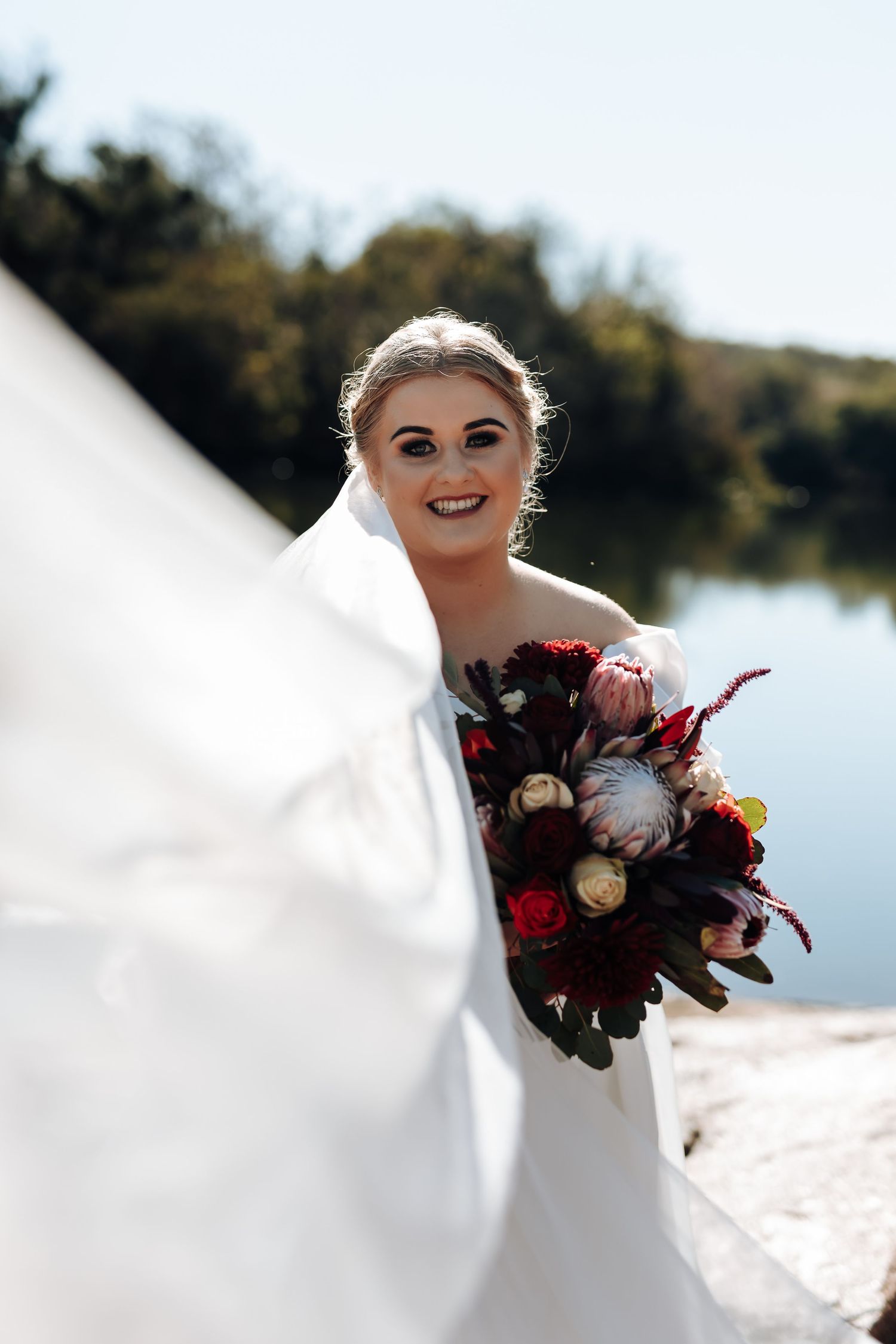 Smiling bride in white dress holds a burgundy and pink bouquet by a sunlit lake, with her veil flowing in the breeze.