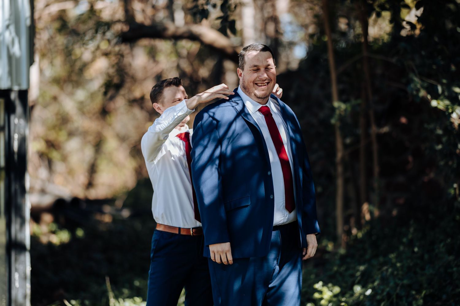 Groomsman helping groom put on blue suit jacket outdoors before wedding ceremony in wooded setting.