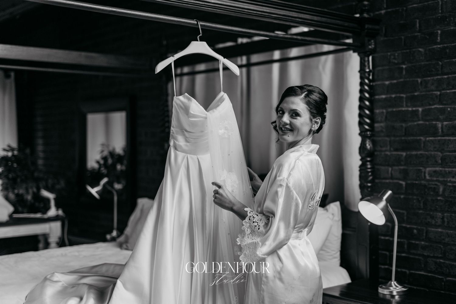Bride in white robe smiling at her wedding dress hanging in a black and white bridal preparation photo.