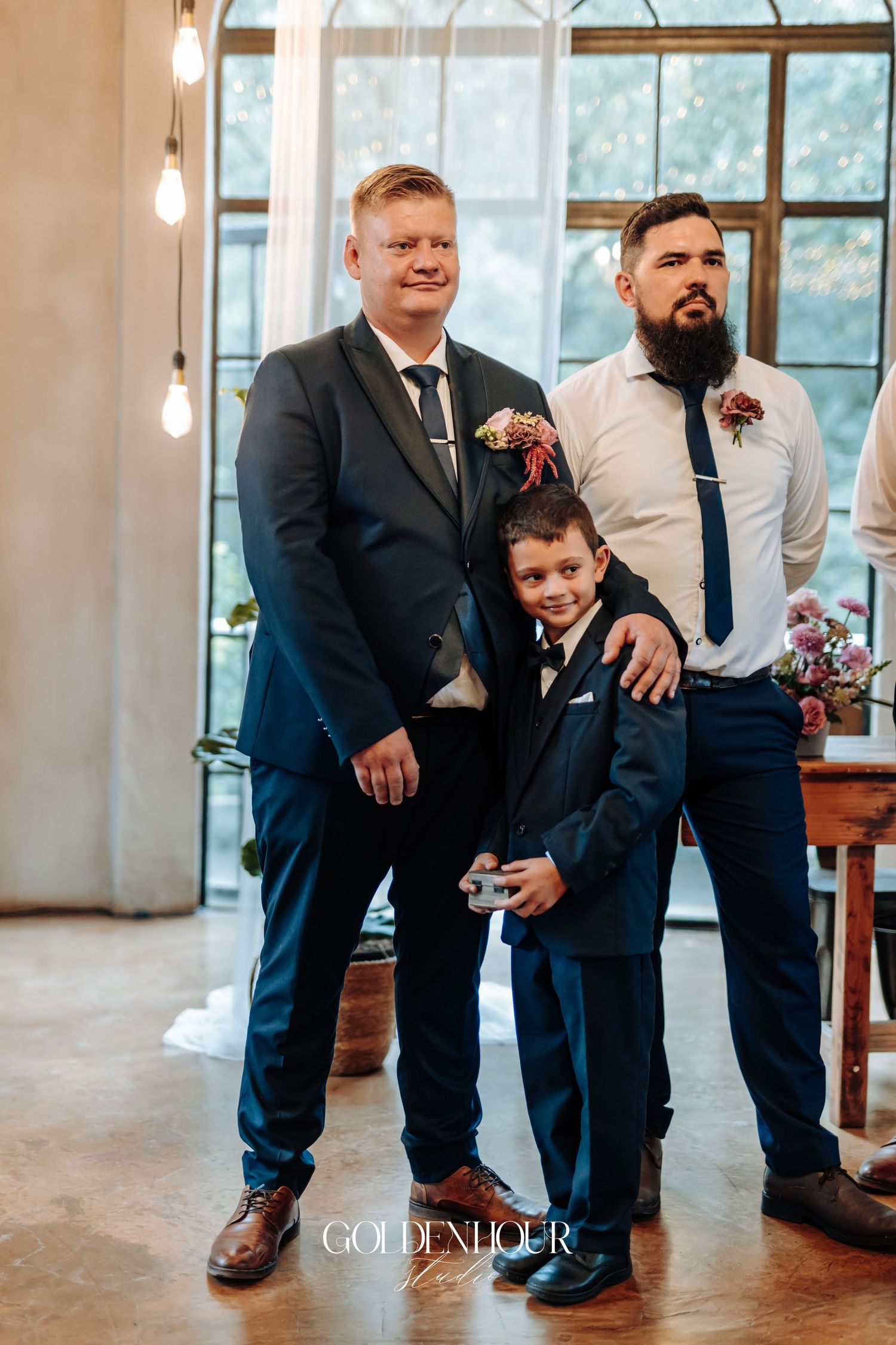 Groom in dark suit standing with bearded groomsman and young boy in navy suit at wedding.