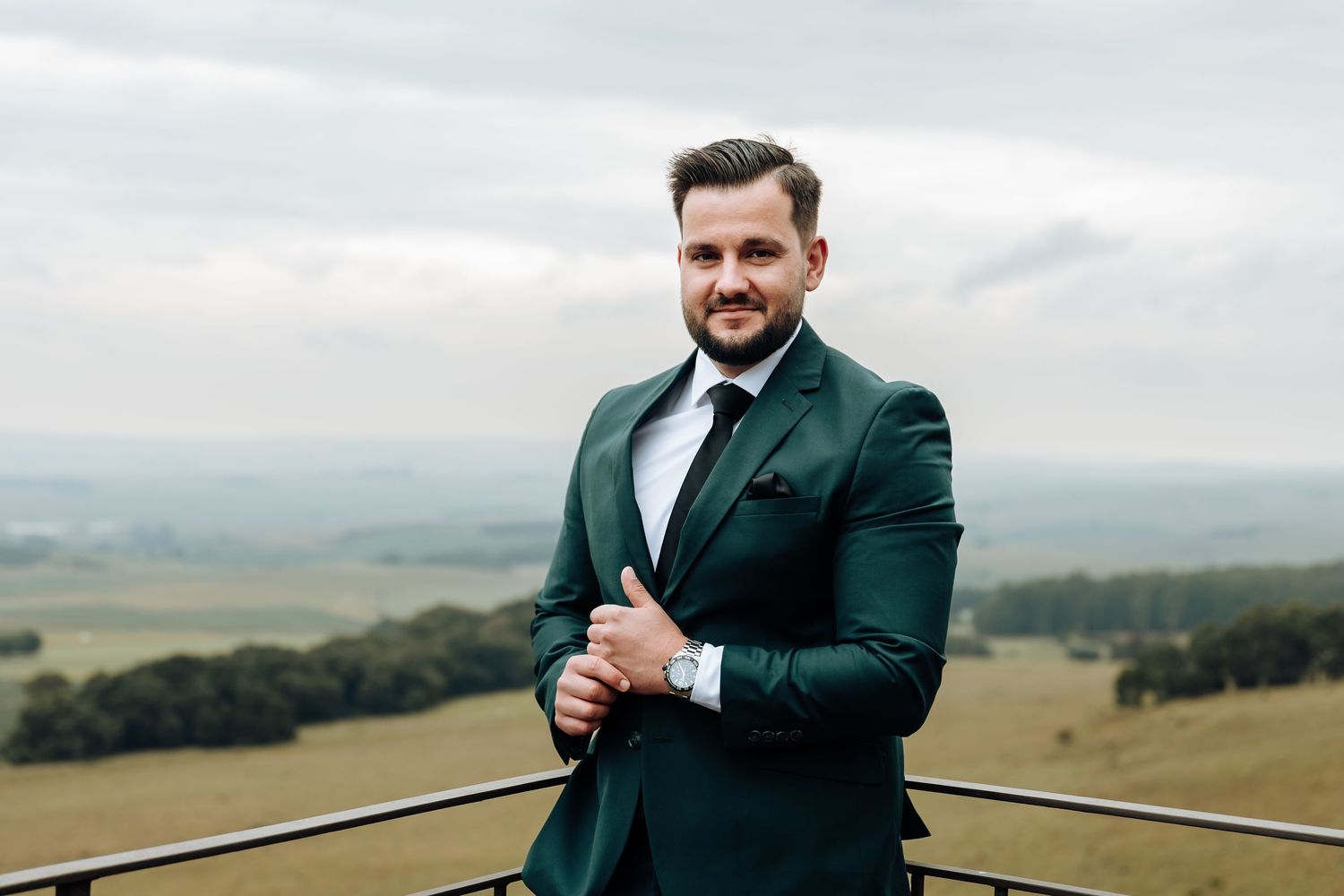 A groom in a dark green suit poses for wedding photos in a scenic outdoor setting with rolling hills in the background.
