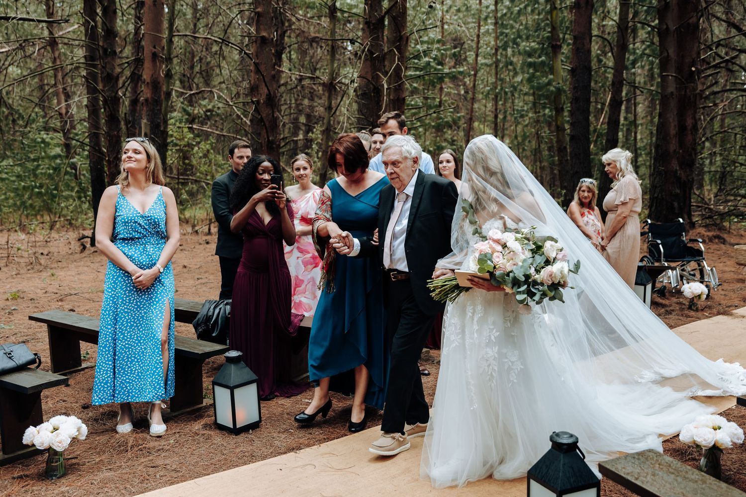 A wedding ceremony takes place in a serene forest setting with guests gathered among tall pine trees.