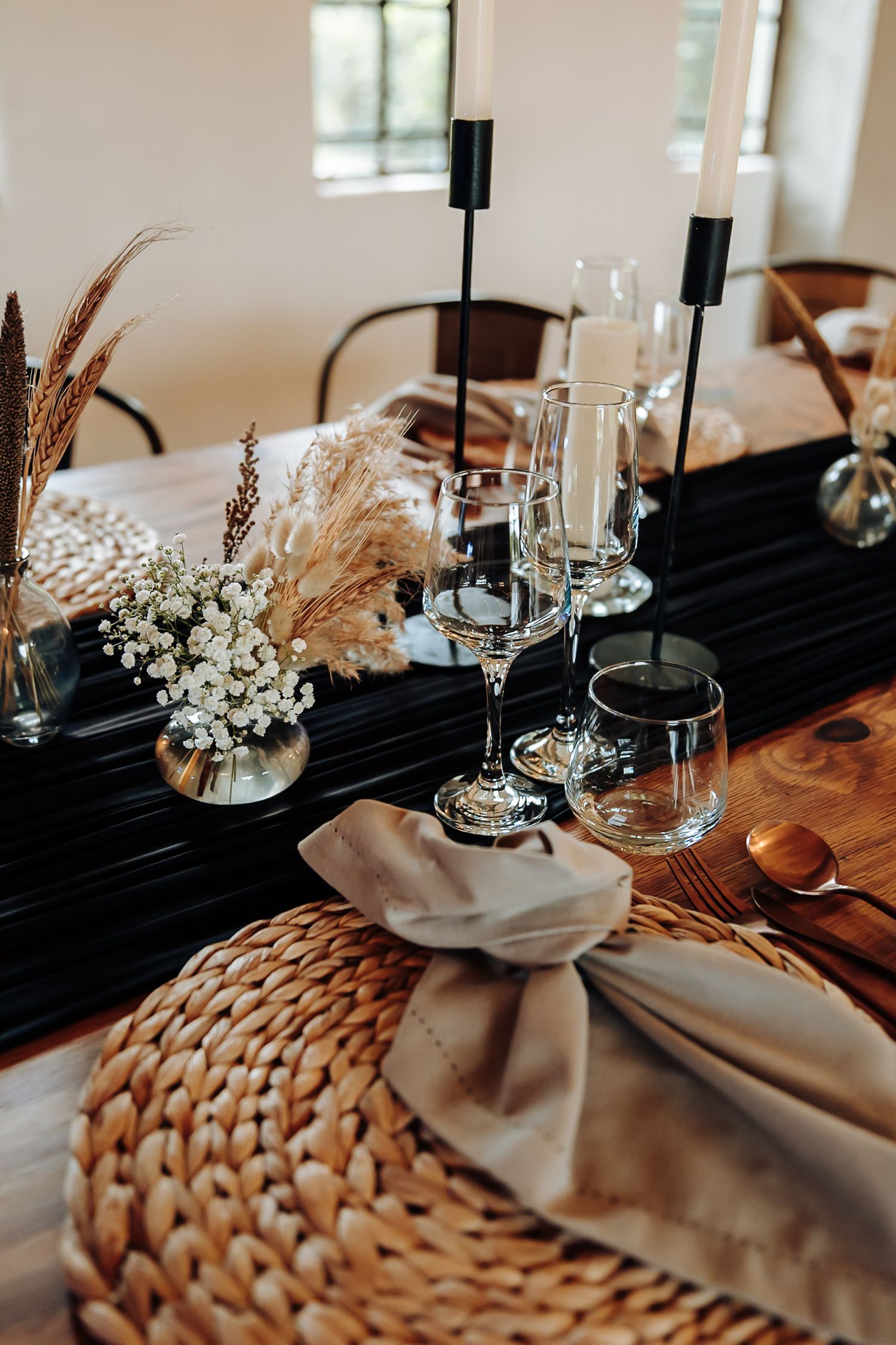 Elegant table setting with black runner, woven placemats, white flowers and pampas grass in modern rustic style.