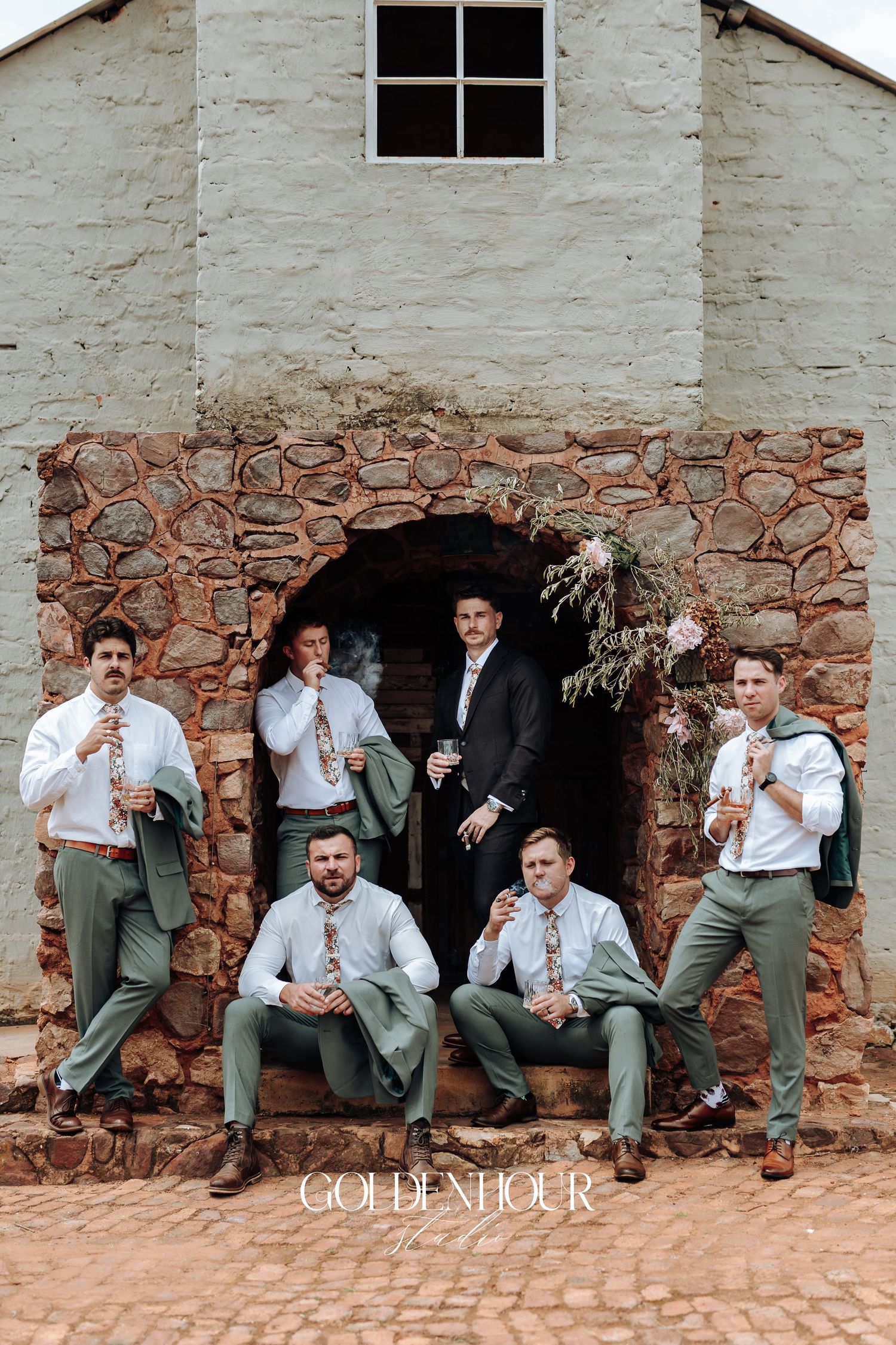 Groomsmen in gray suits pose casually against a rustic stone wall for a fun wedding photo series.