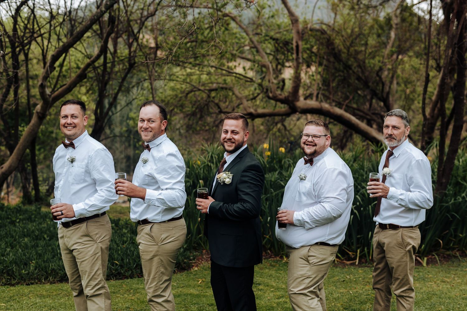 Wedding party in coordinated attire stands together holding drinks in a wooded outdoor setting.