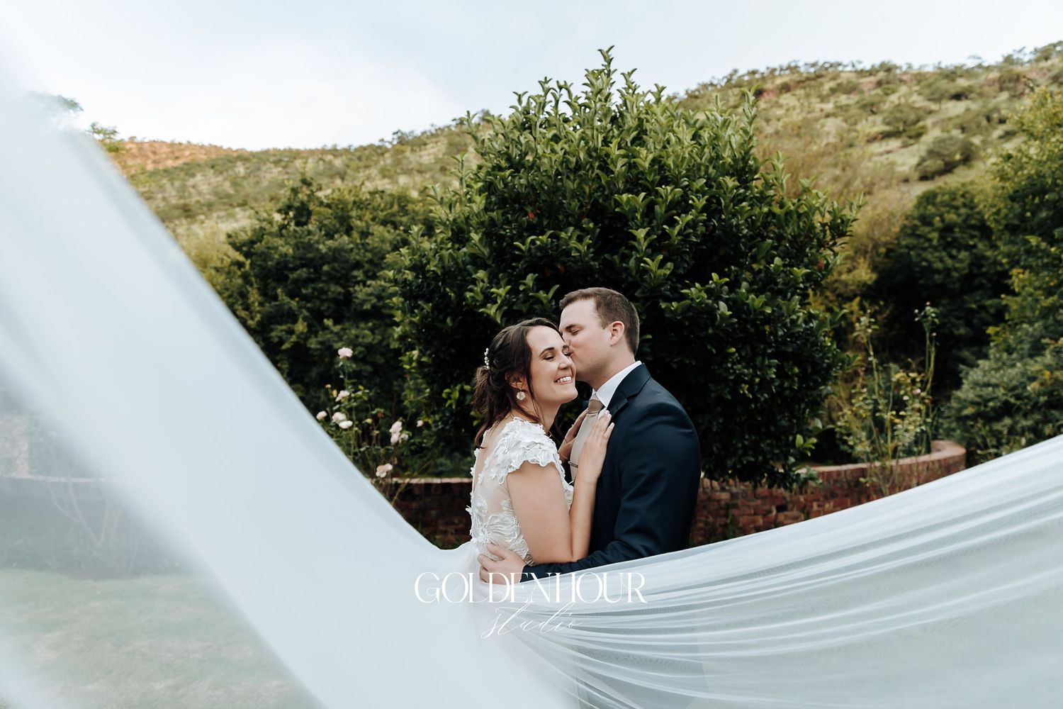 Bride and groom share a romantic embrace outdoors with veil flowing in the breeze and lush green hills behind them.