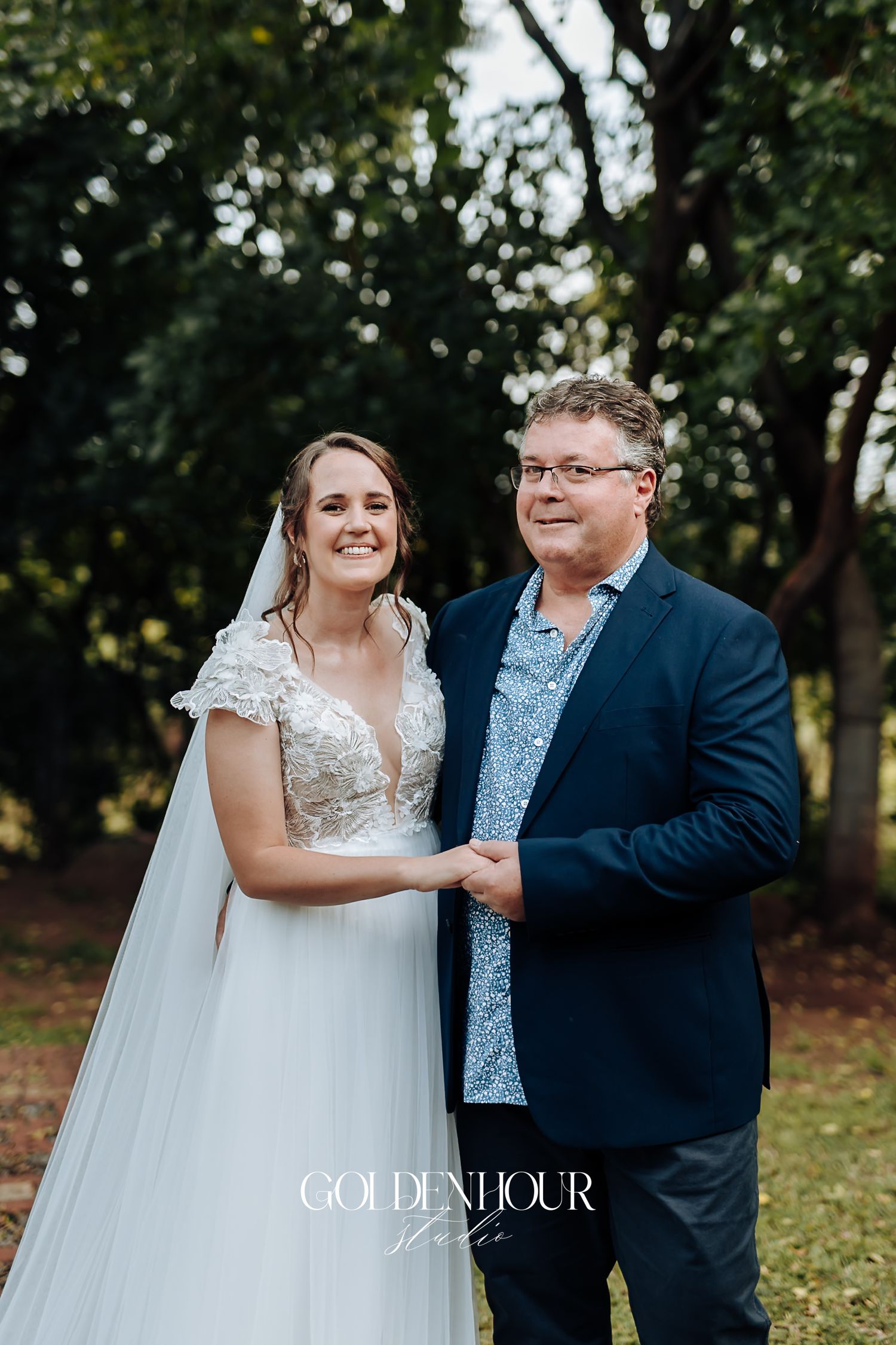 Smiling bride in lace wedding dress holding hands with older man in navy suit outdoors.
