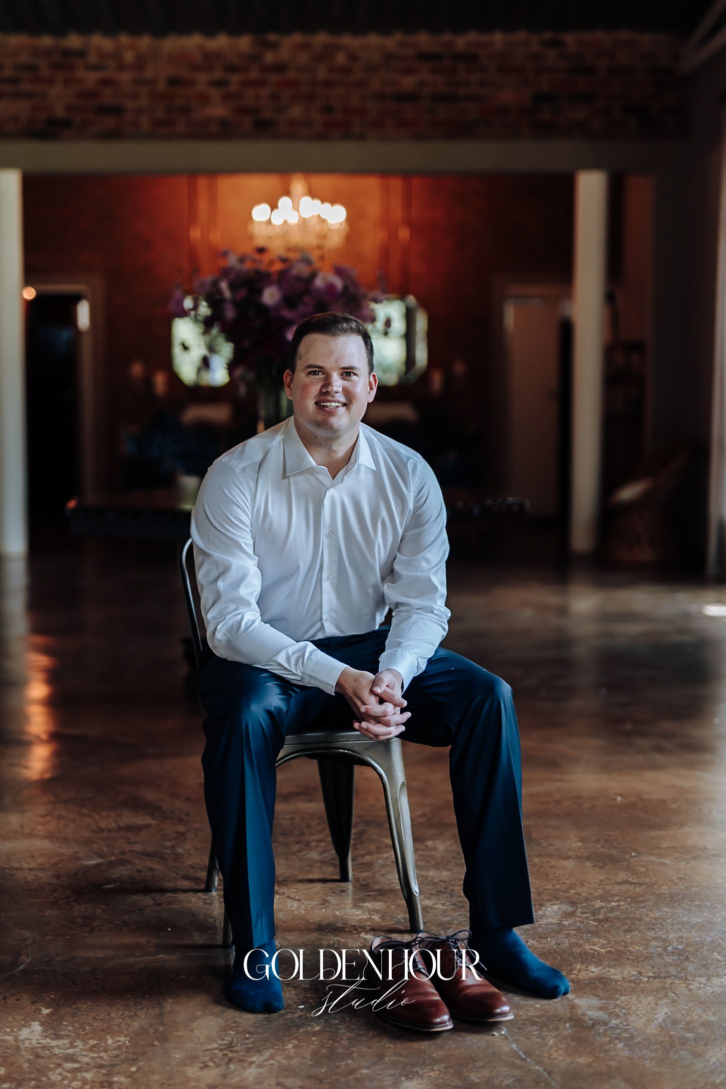 Groom in white shirt and navy pants sits smiling on a metal chair in an elegant wedding venue.