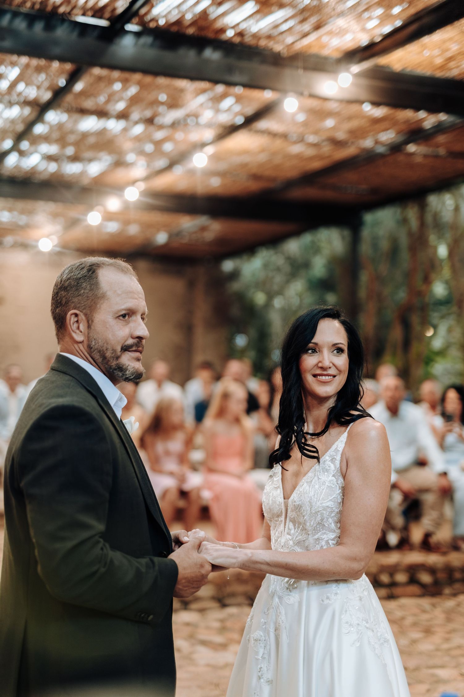 Bride and groom holding hands at outdoor wedding ceremony under rustic pergola with string lights and guests seated behind.