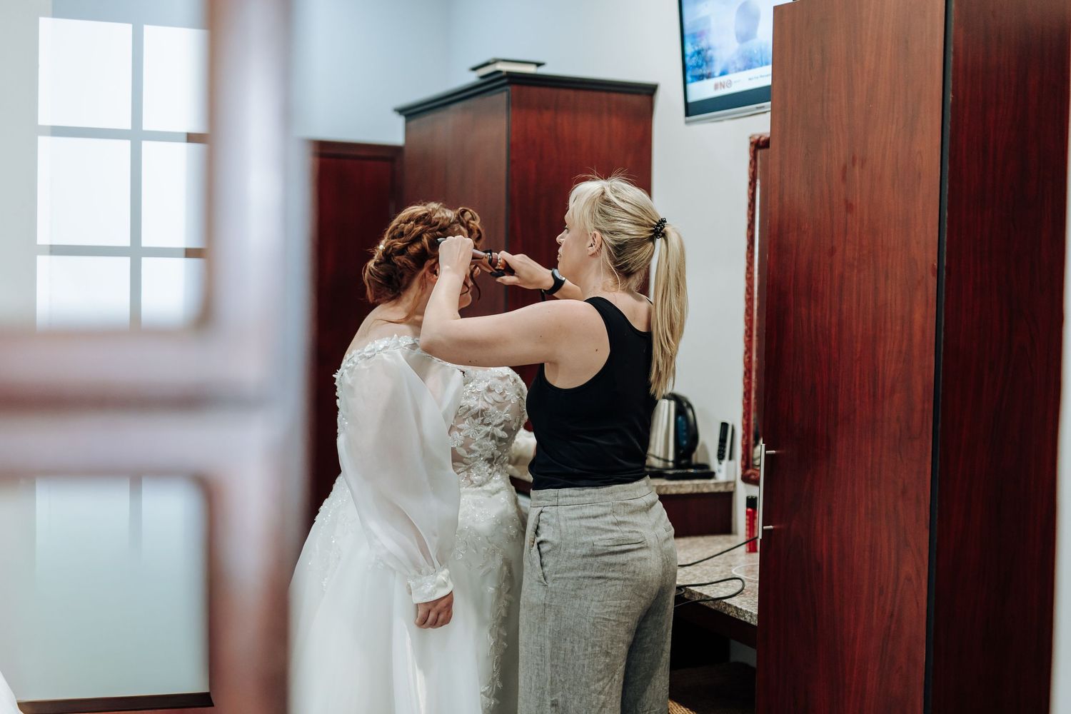 A hairstylist works on styling a bride's hair in a white wedding dress during pre-wedding preparations.