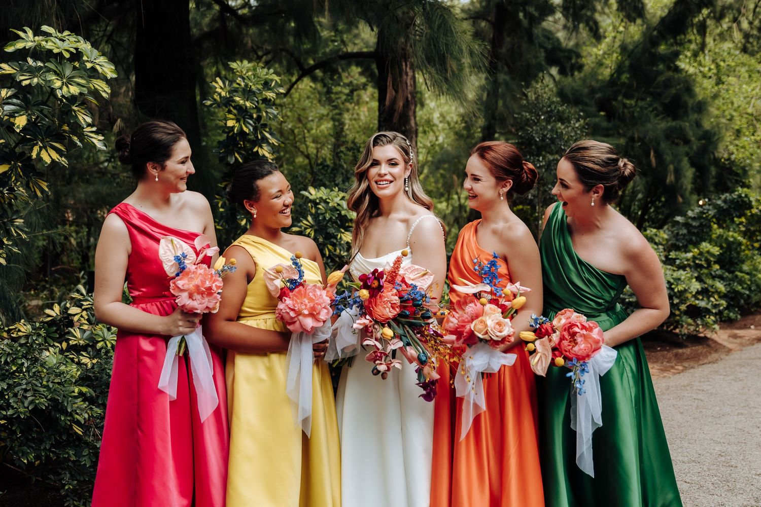 Bridesmaids in colorful rainbow dresses from red to green stand together holding coral bouquets in an outdoor setting.