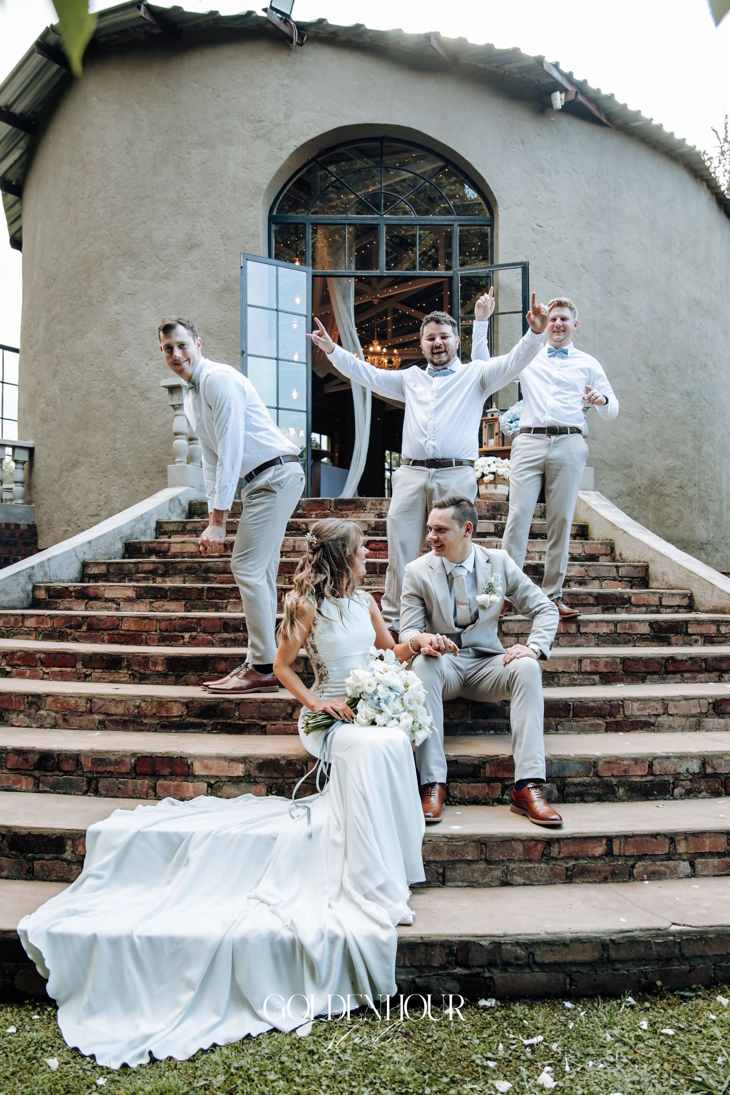 Playful wedding couple poses on rustic brick steps outside a stucco building with creative dance-inspired movements.