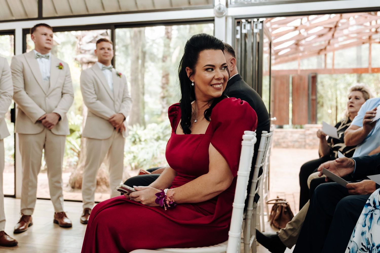 Wedding guests in tan suits stand near someone in a burgundy dress seated in a chair at an indoor venue.