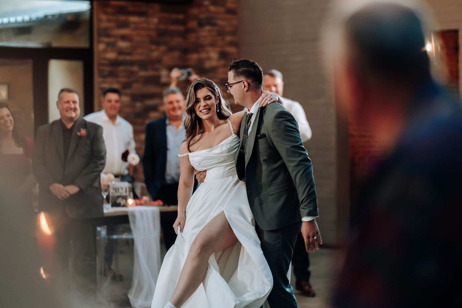 A couple dances together at their wedding reception in front of guests against a brick wall backdrop.