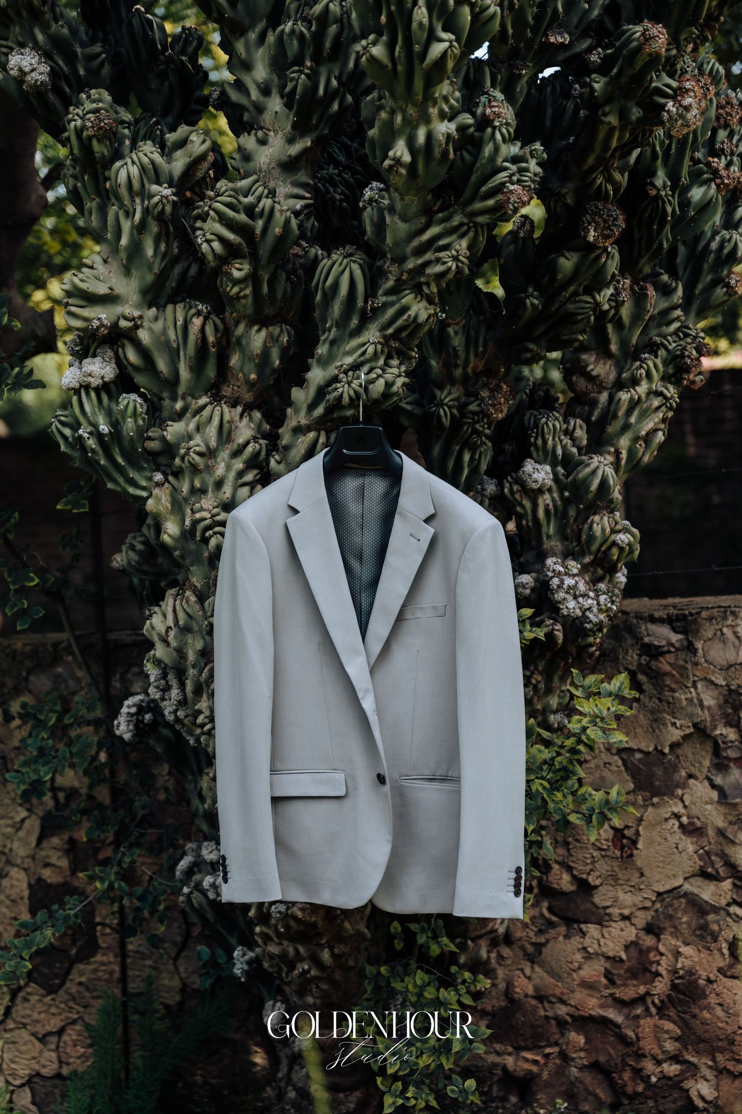 A light gray suit jacket hangs elegantly against a lush green garden backdrop with stone wall details.