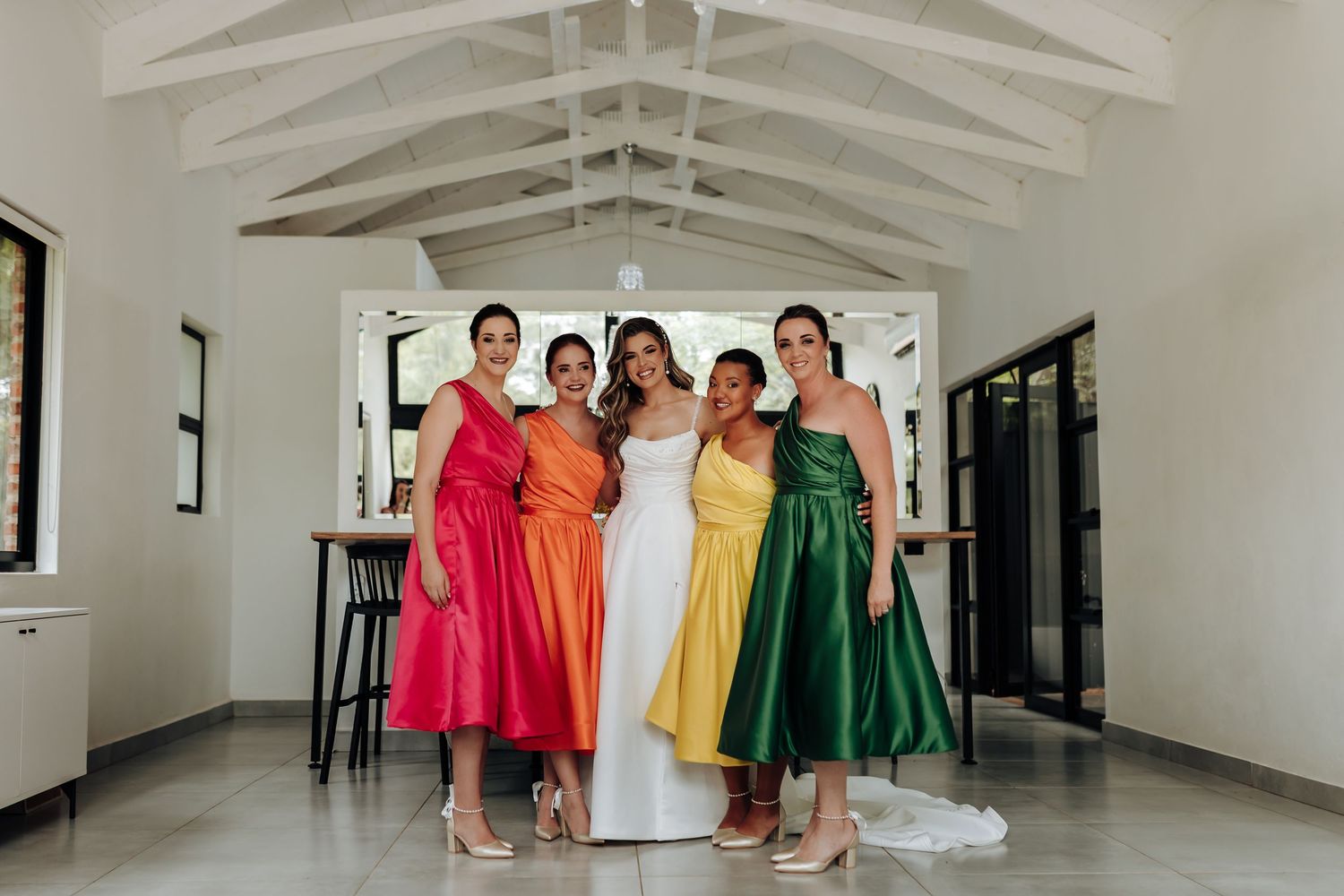 Wedding party poses in colorful tea-length dresses inside bright modern venue with vaulted ceilings.