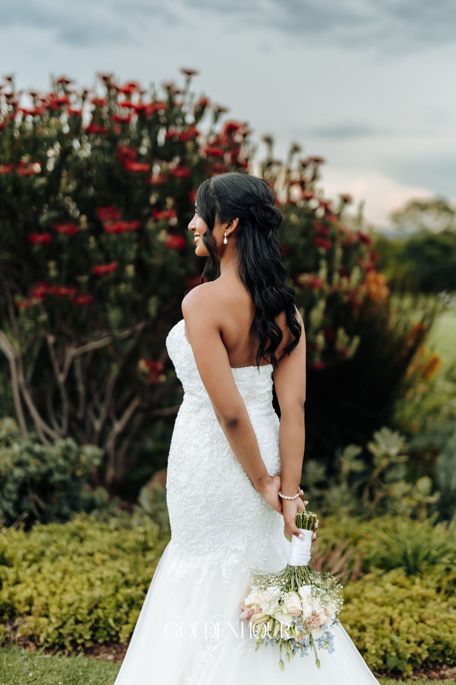 Bride in white lace gown holding bouquet near blooming red flowers.
