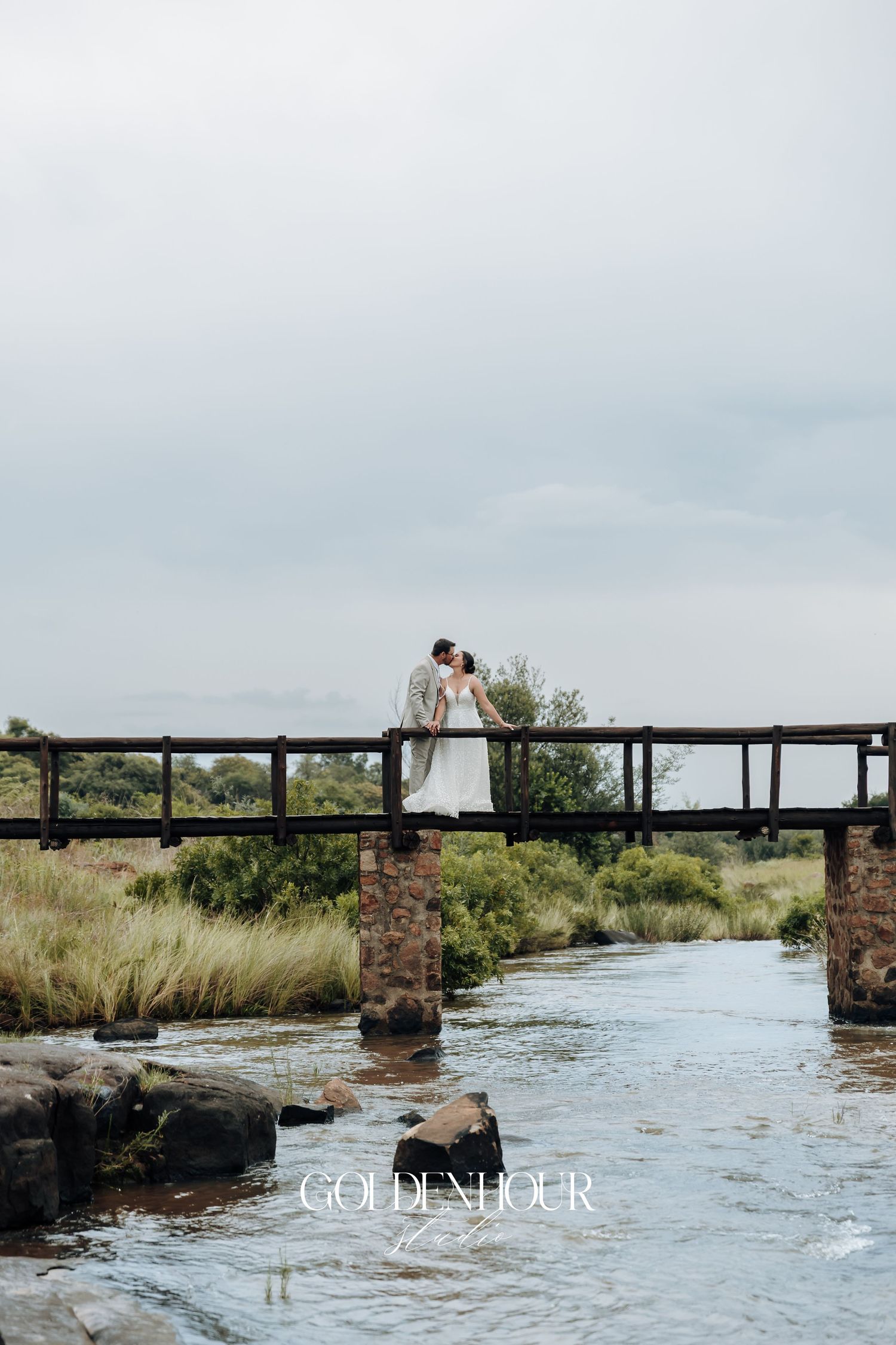 Wedding couple poses on rustic bridge over flowing river with scenic landscape views.